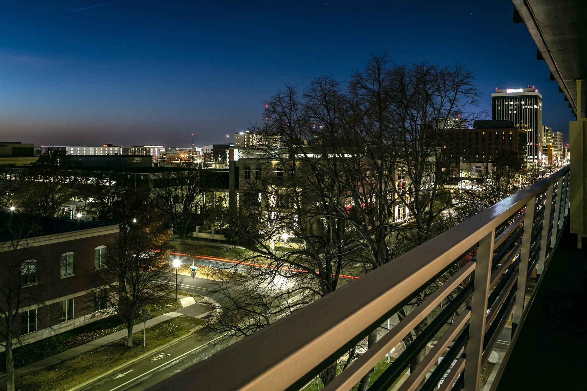 Night view of a city courtyard with lit buildings, trees, and a balcony railing in the foreground