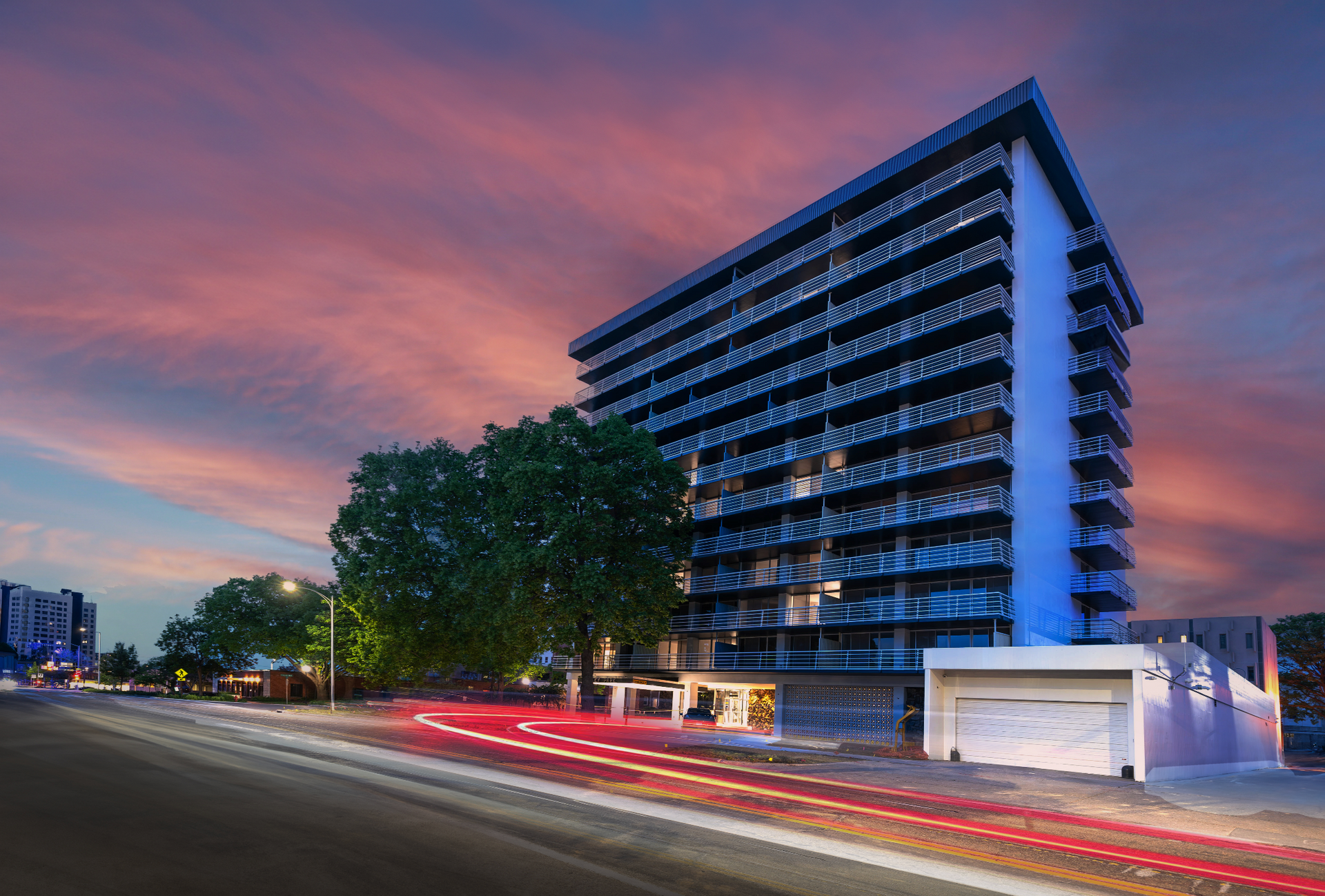Modern apartment building at dusk with pink sky and red car light trails on a city street