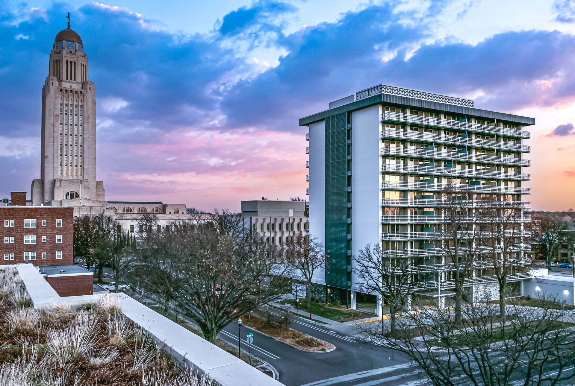 City skyline at dusk with lit buildings, trees, and a balcony railing in the foreground