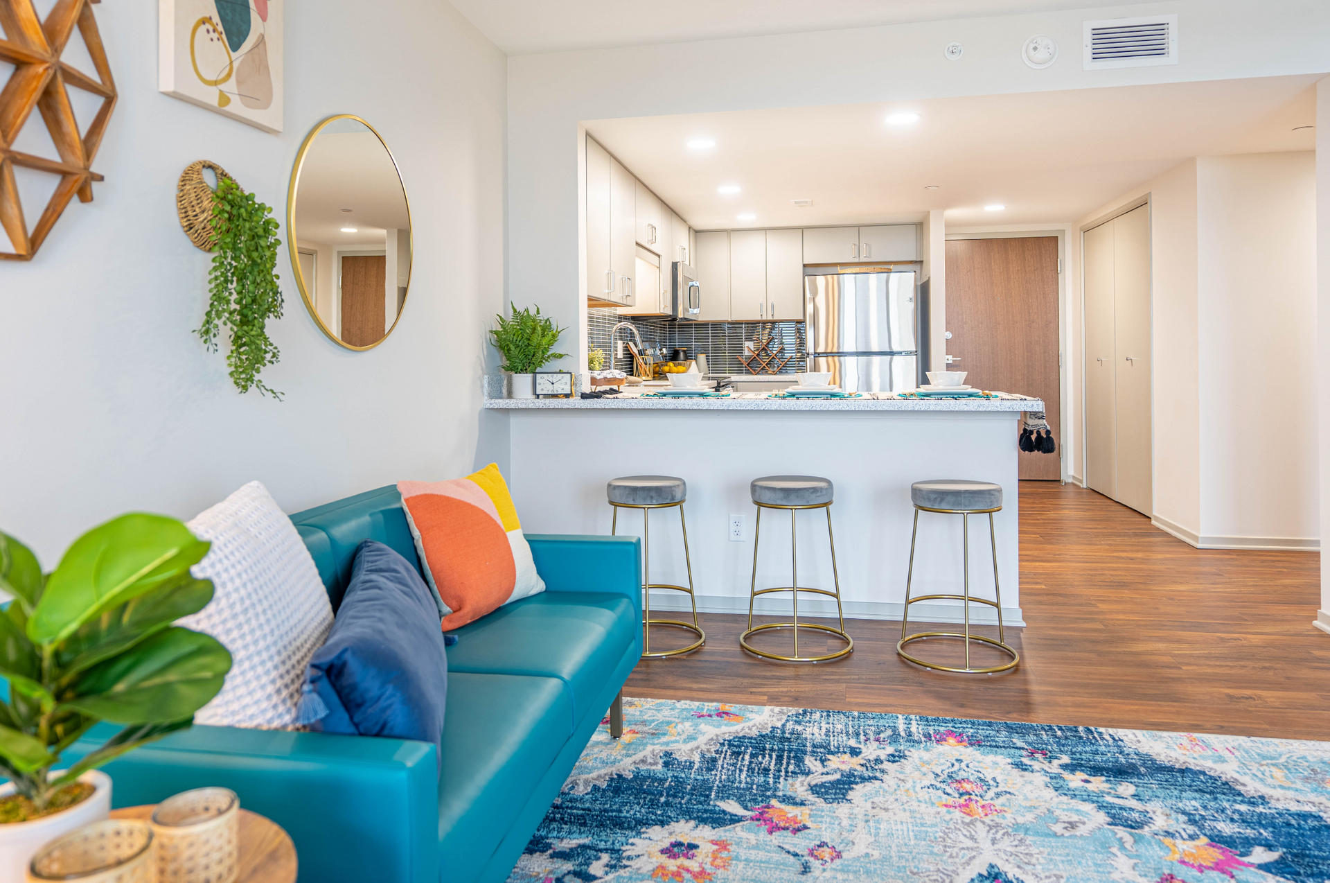 Bright modern living room with teal sofa, colorful rug, and kitchen bar stools in the background