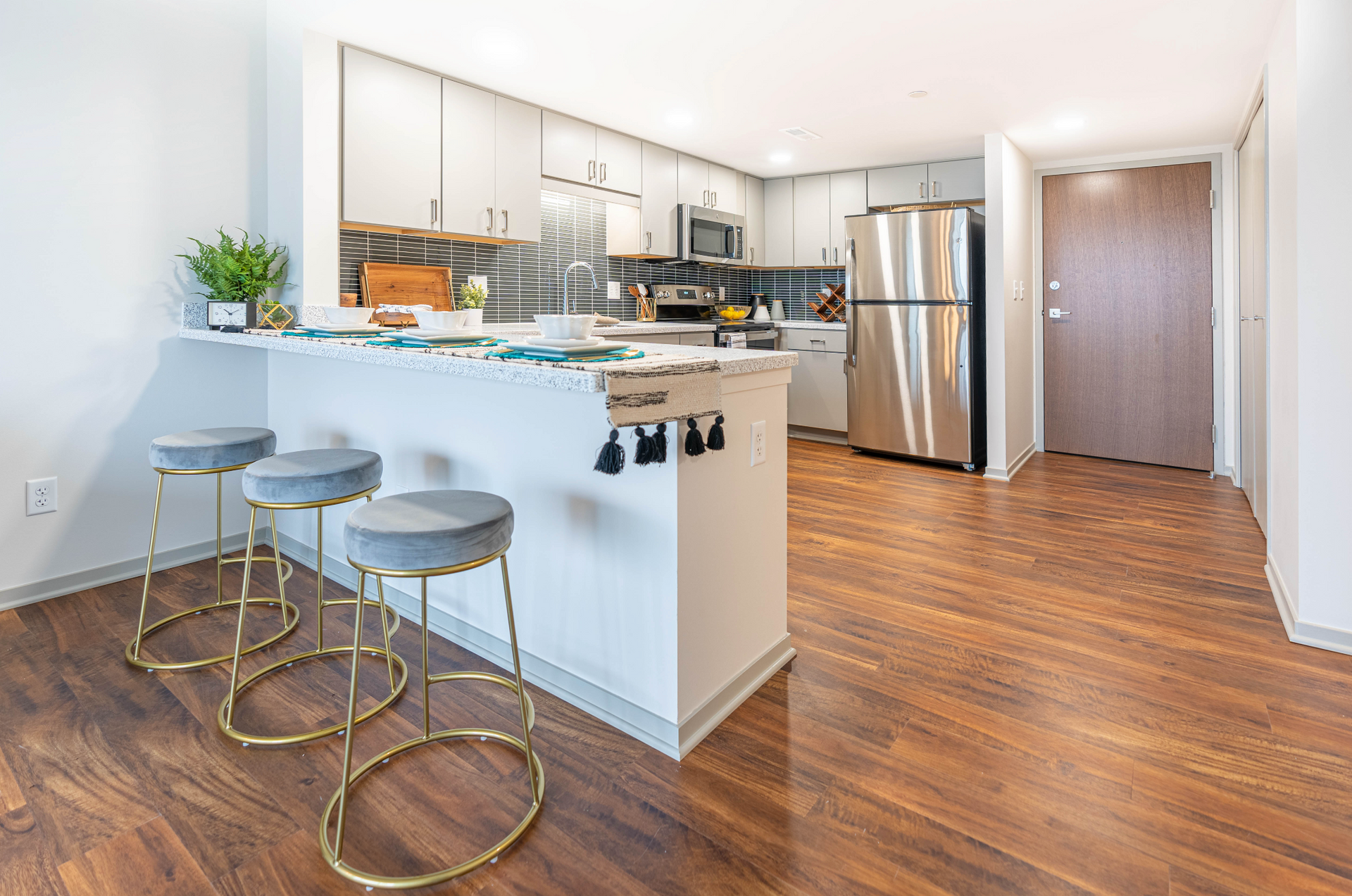 Modern kitchen with white cabinets, breakfast bar, gold stools, stainless appliances, and wood floors.