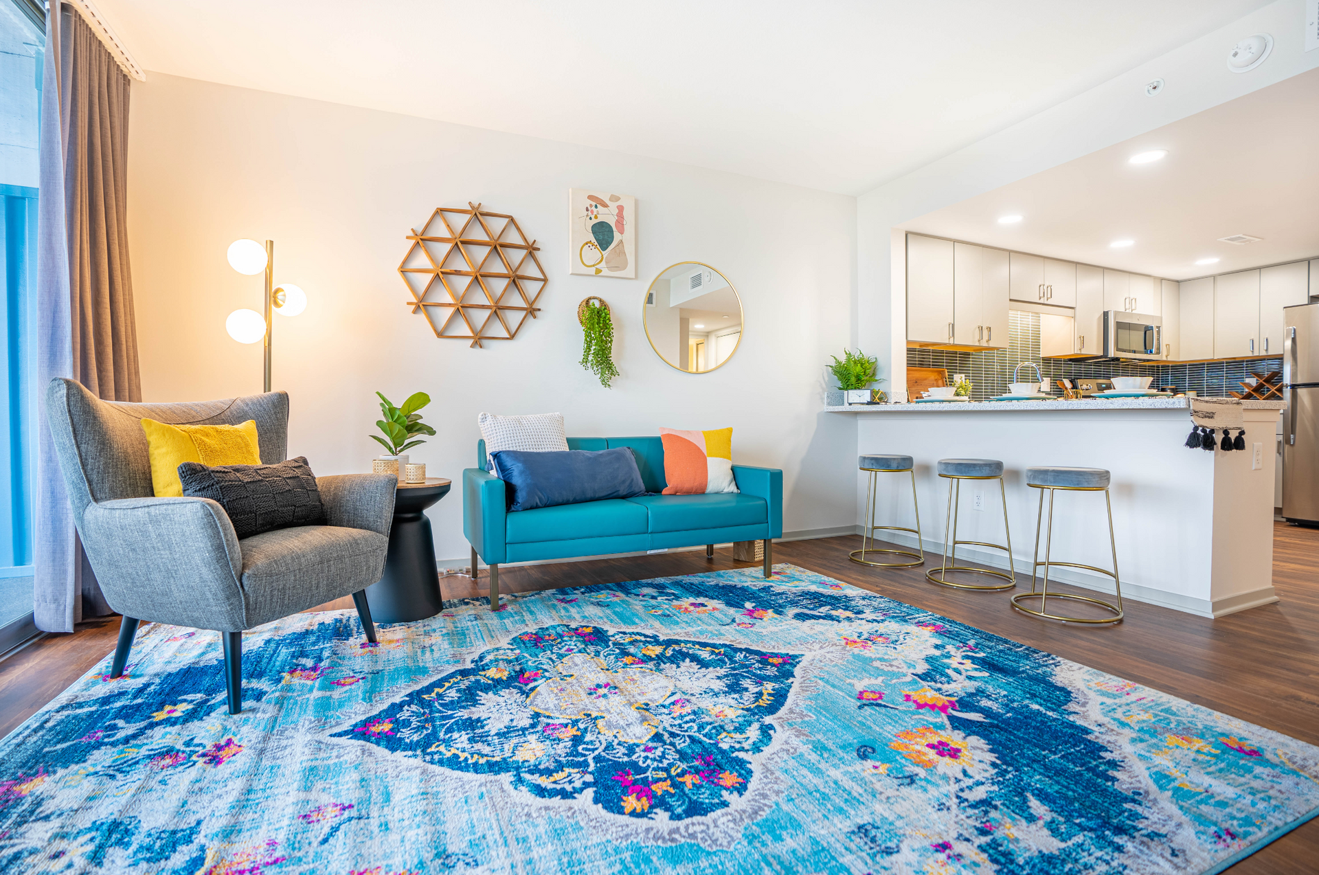 Bright living room with colorful rug, teal sofa, armchair, wall decor, and open kitchen bar stools