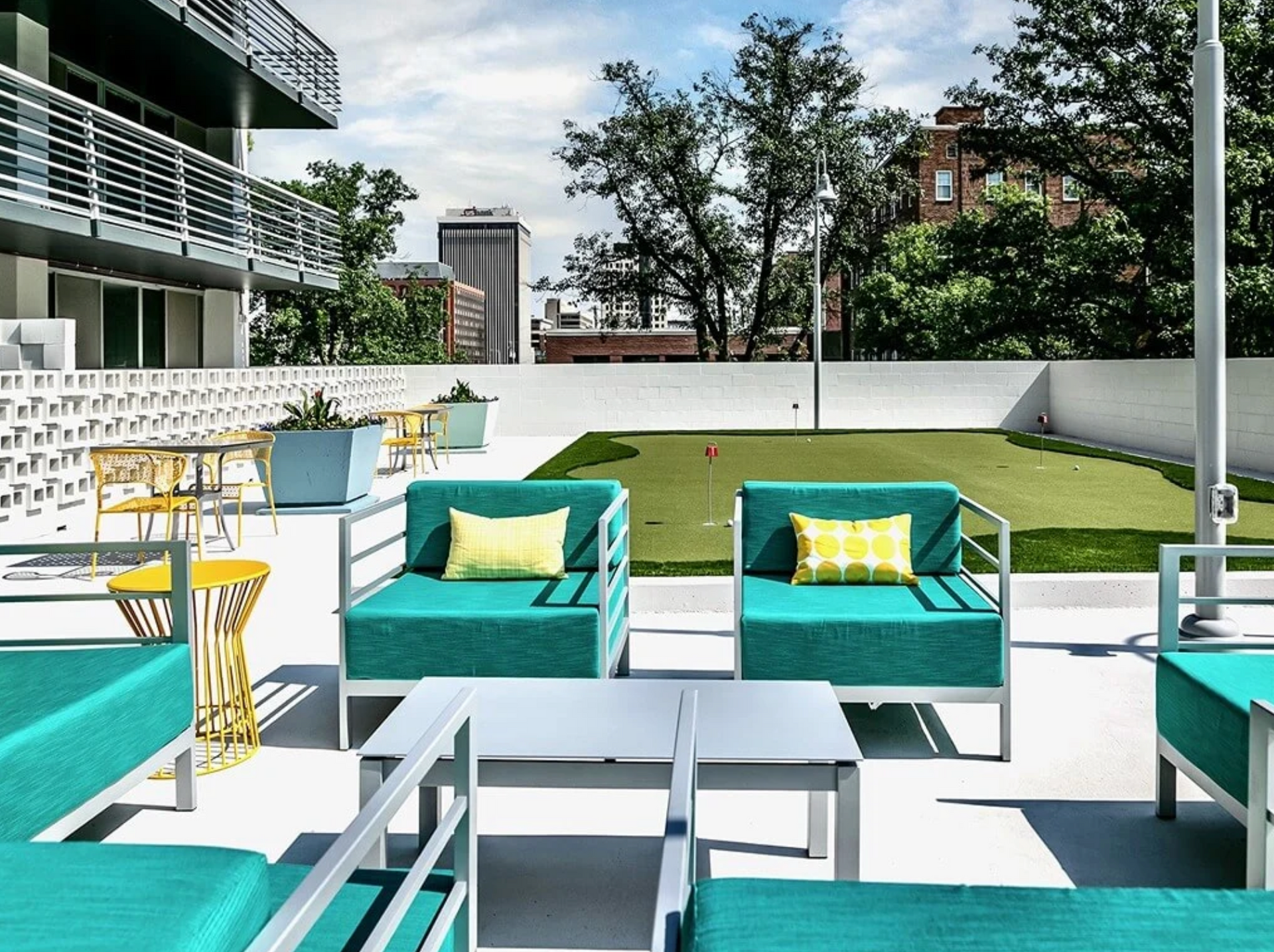 Outdoor rooftop lounge with turquoise chairs, white tables, yellow pillows, and a grassy area beside a modern building