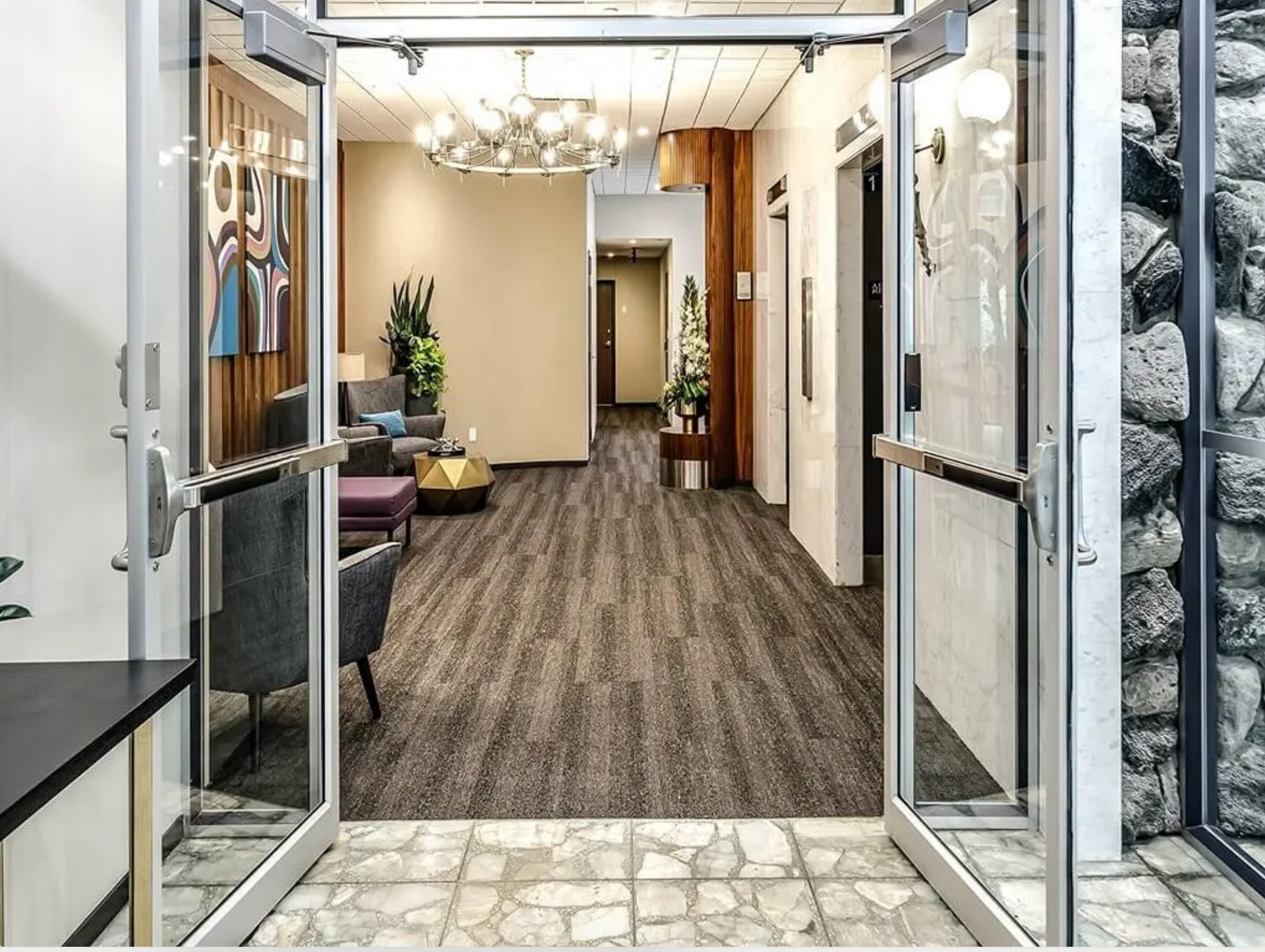Open glass doors lead into a modern lobby with wood flooring, chandelier lighting, and seating.