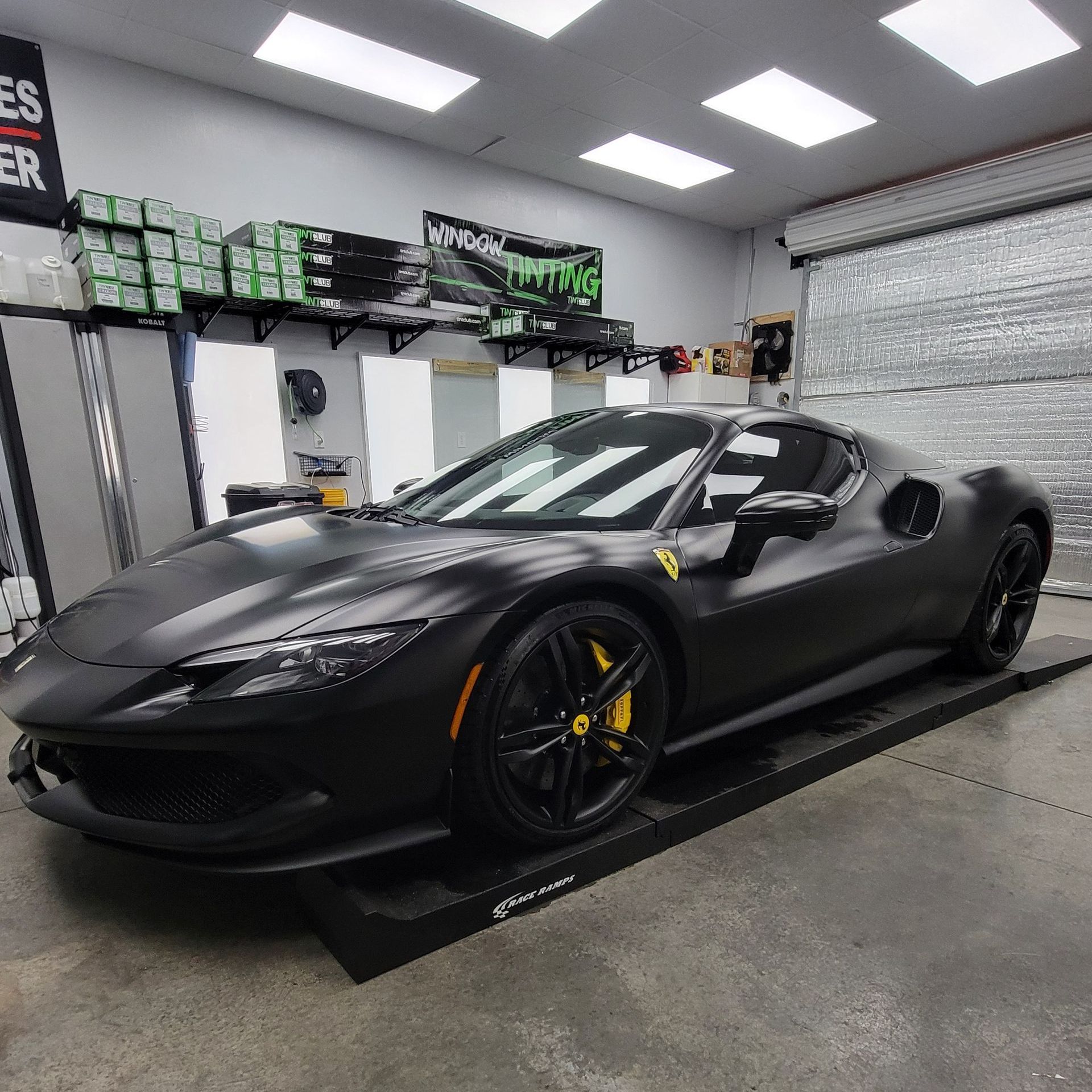 Matte black Ferrari sports car in a garage, with yellow brake calipers and tinted windows.