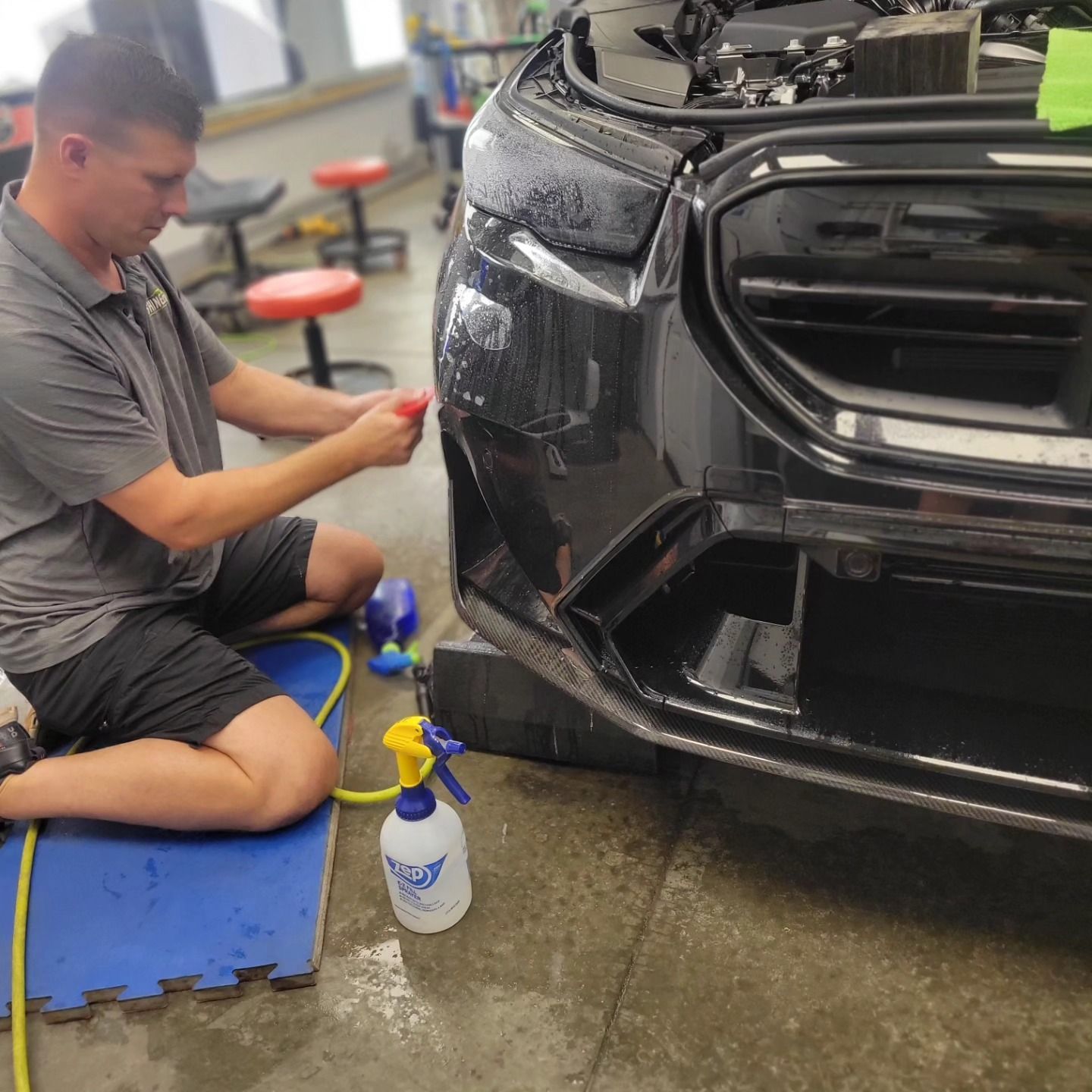 Man applying film to a black car's bumper in a workshop, using spray bottle and tools.