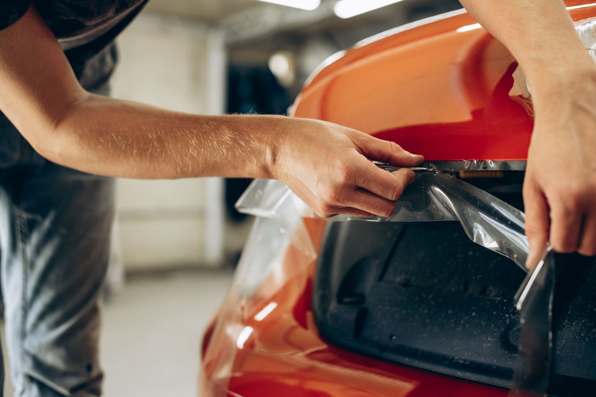 Person applying protective film to a red car bumper in a workshop.
