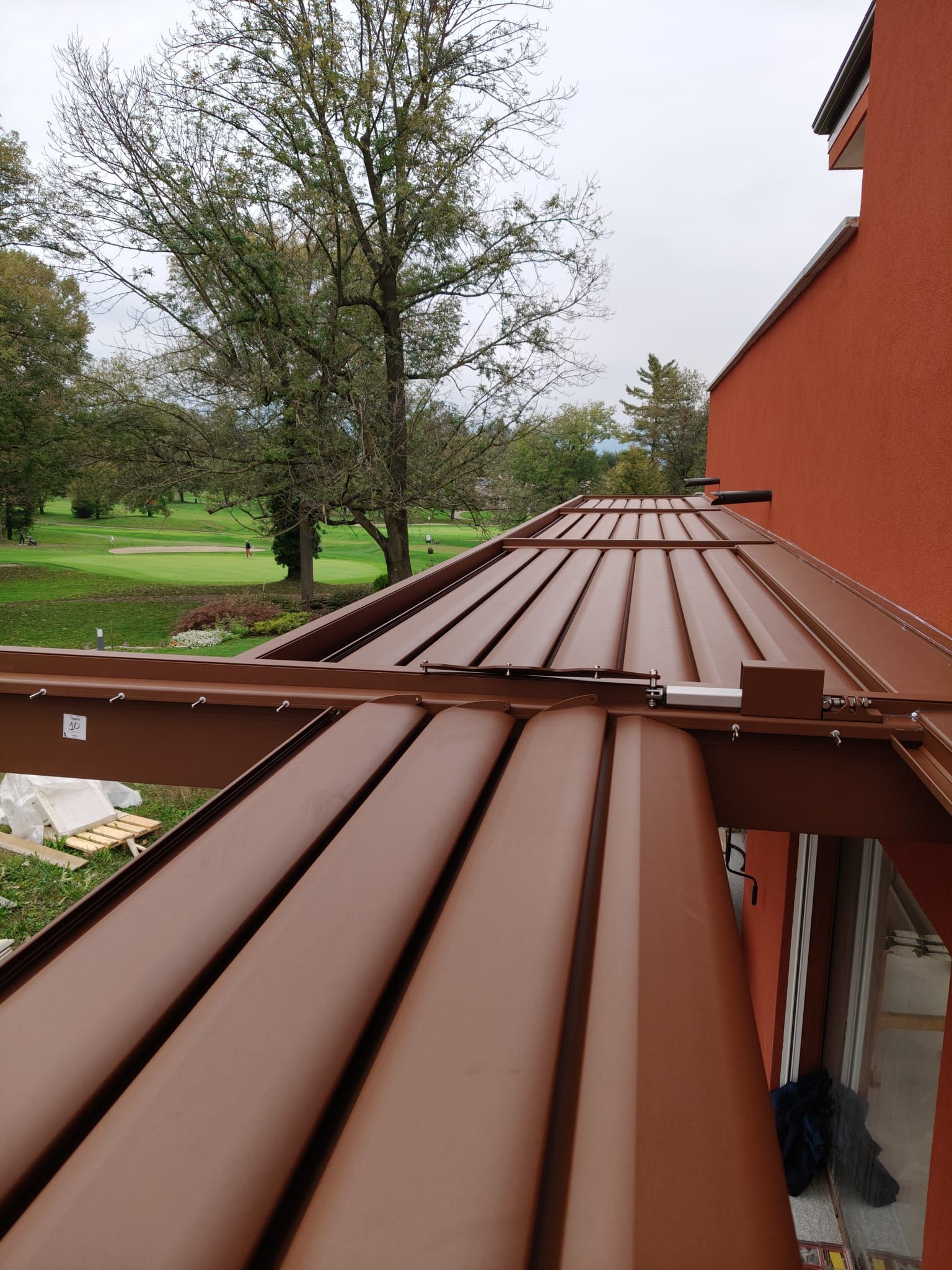 Pergola marrone con stecche parzialmente aperte che si affaccia sul prato verde e sugli alberi.