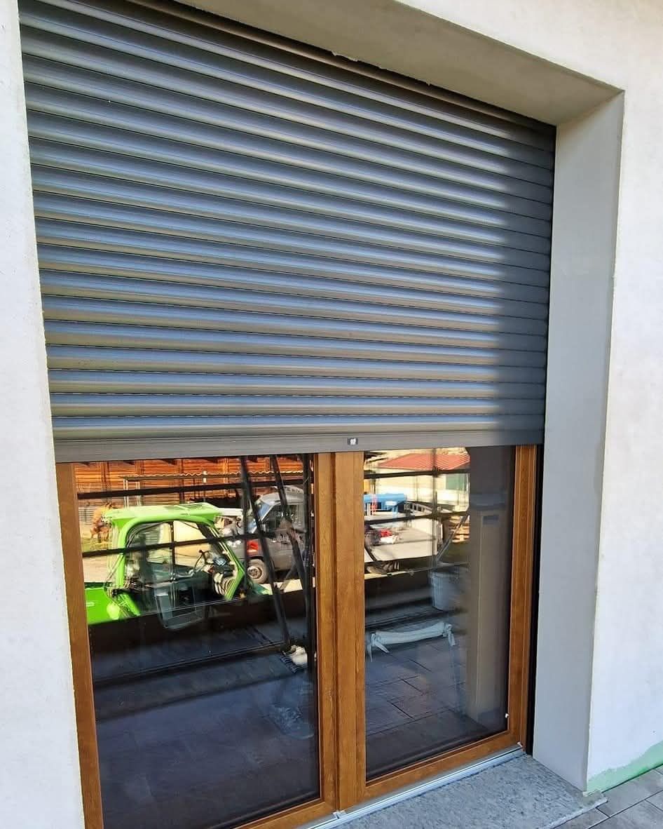 Finestra con tapparella grigia chiusa; telaio in legno, riflesso di un'auto verde brillante.
