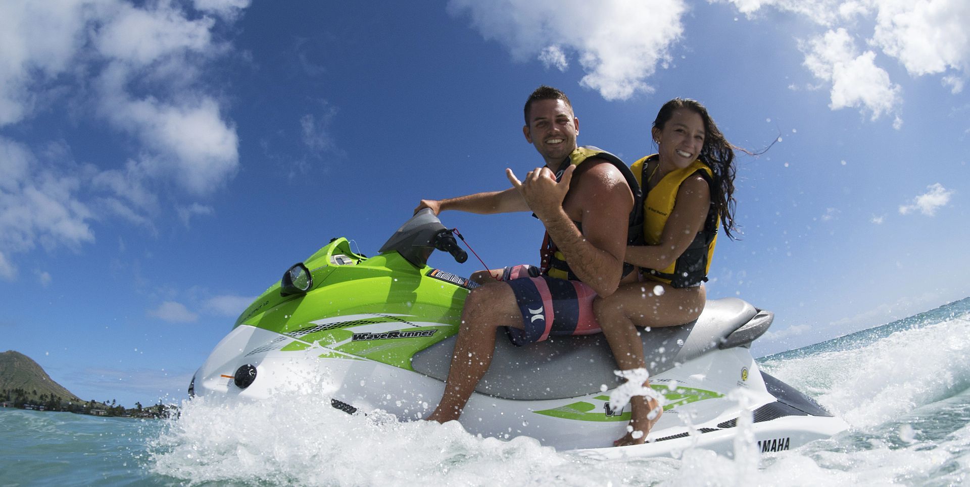 Two people ride a jet ski, splashing through blue water under a bright sky.