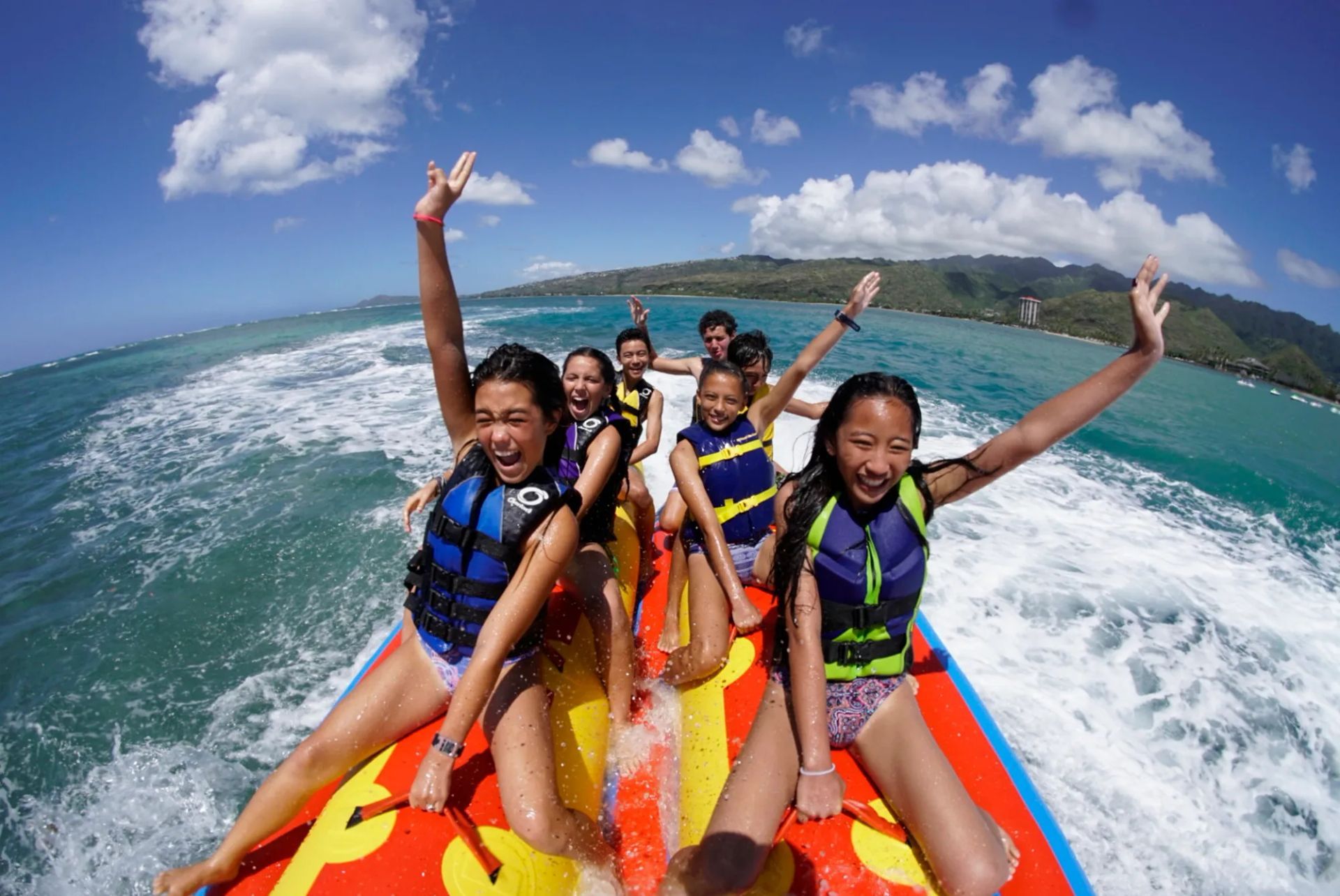 People on a colorful banana boat ride, smiling and raising their arms on a sunny ocean.