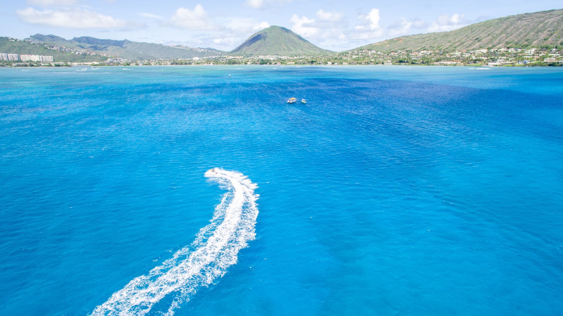 Bright turquoise ocean with a white wake trailing from a boat, mountains and blue sky in the distance
