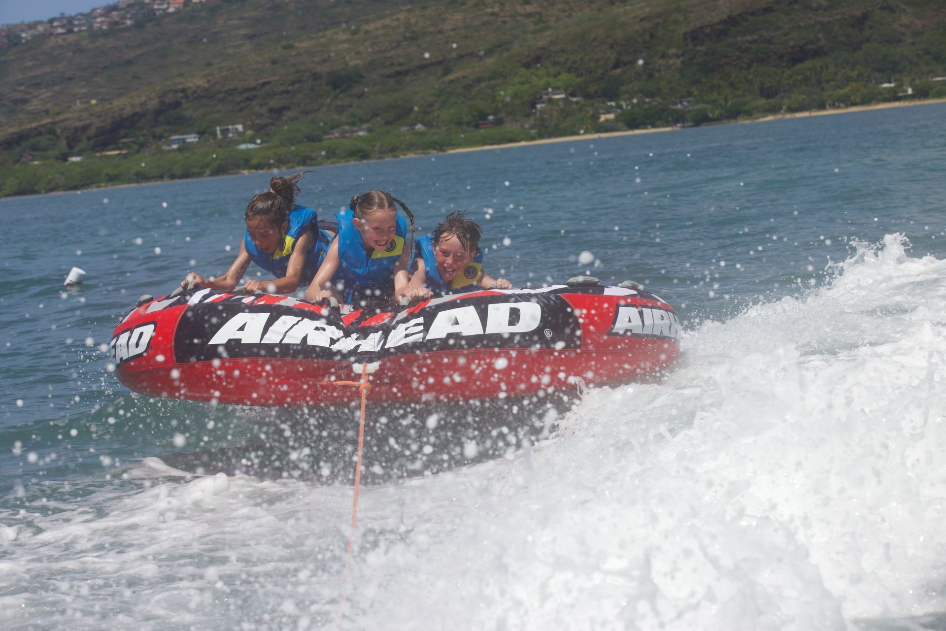 People riding a red bumper tube through water, splashing near a green shoreline