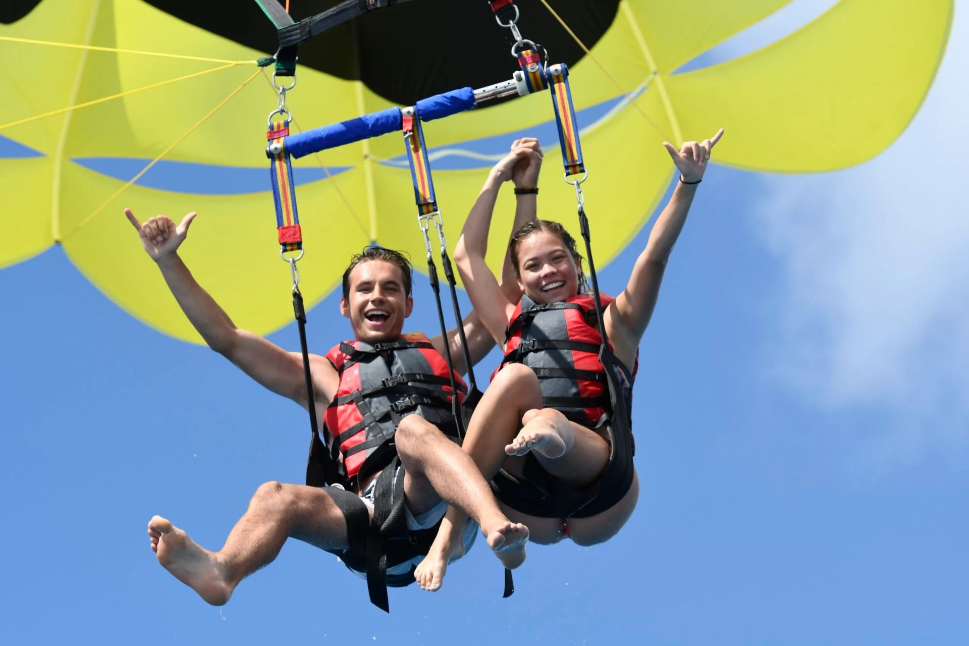 Two people parasailing with red harnesses against a blue sky and bright yellow canopy