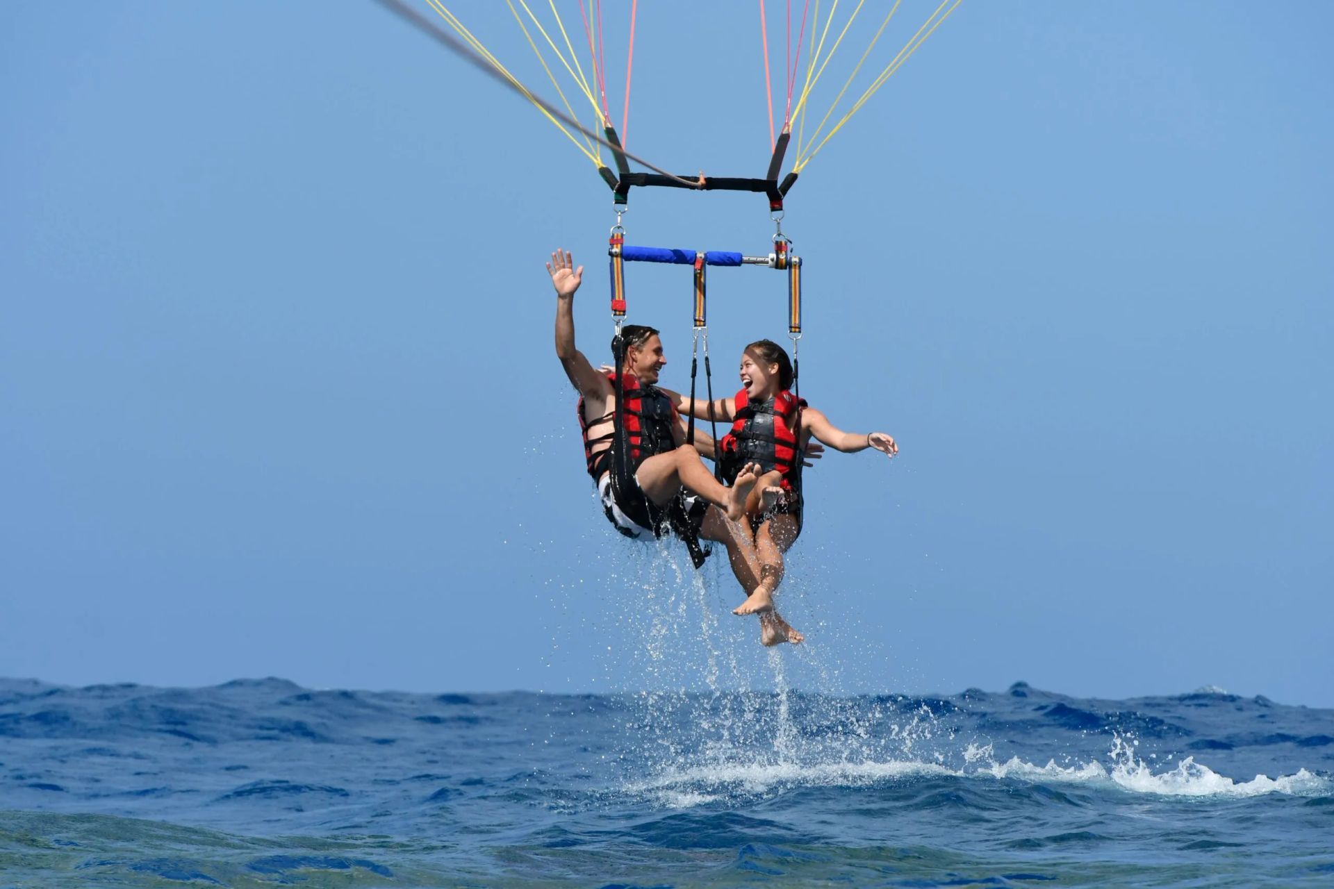 Two people parasailing over the ocean, suspended above blue water under a clear sky