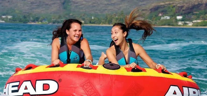 Two people laughing on a bumper tube being towed across blue water
