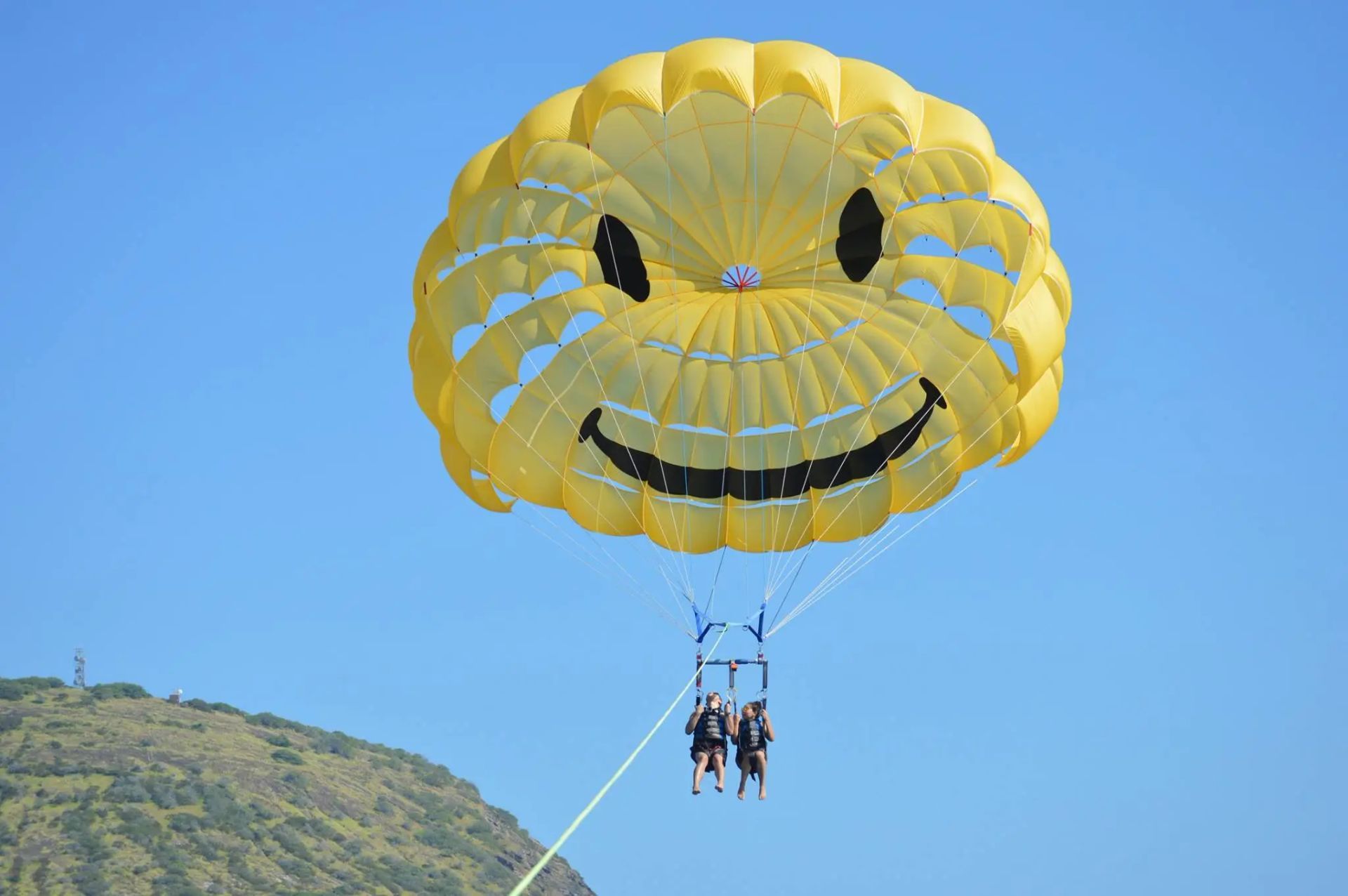 Bright yellow smiley-face parasail carrying two people over the ocean near a green hillside