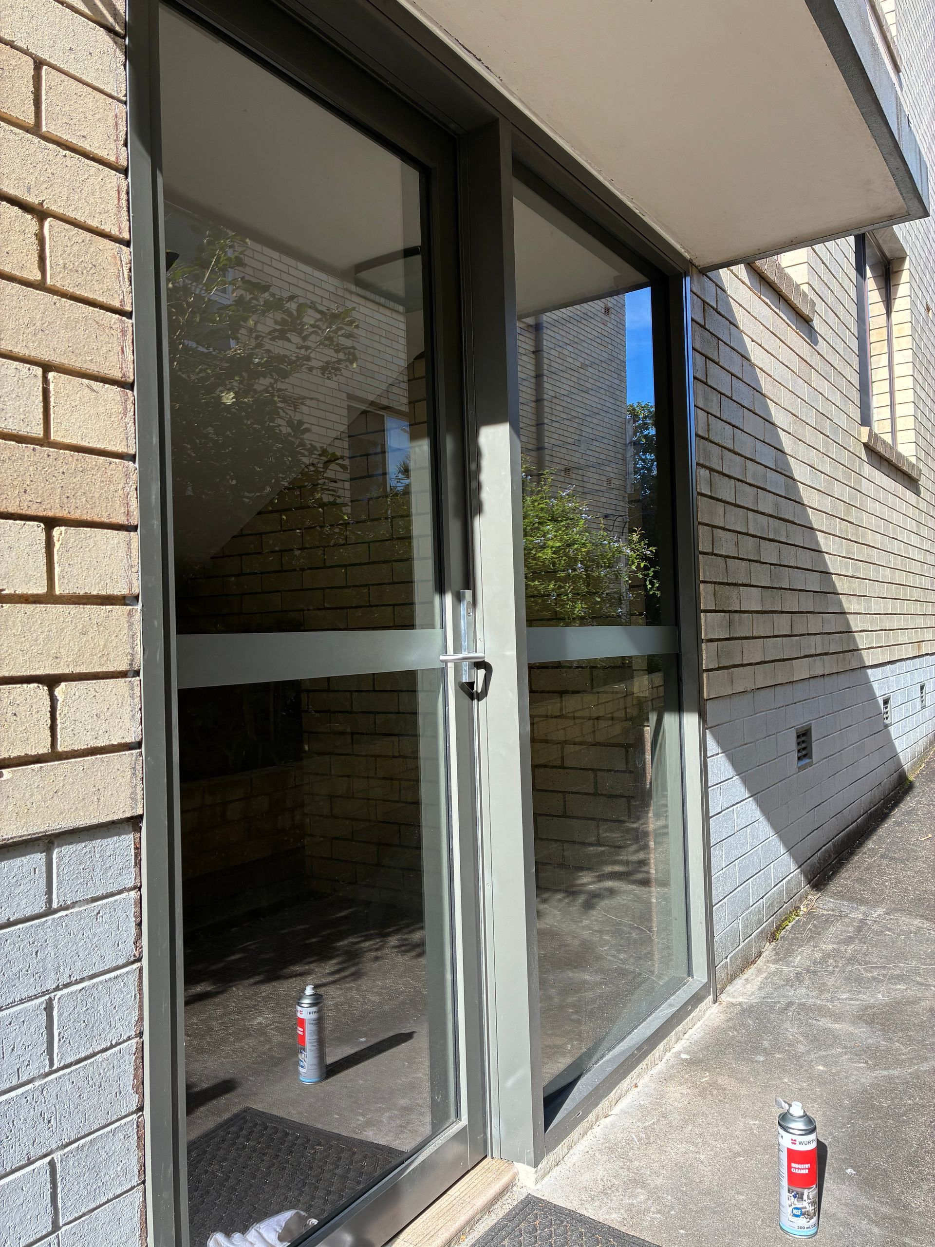 Glass entrance doors in a brick building exterior, with sunlight and two red cans on the ground. — Kracken Windows and Doors In Terrey Hills, NSW