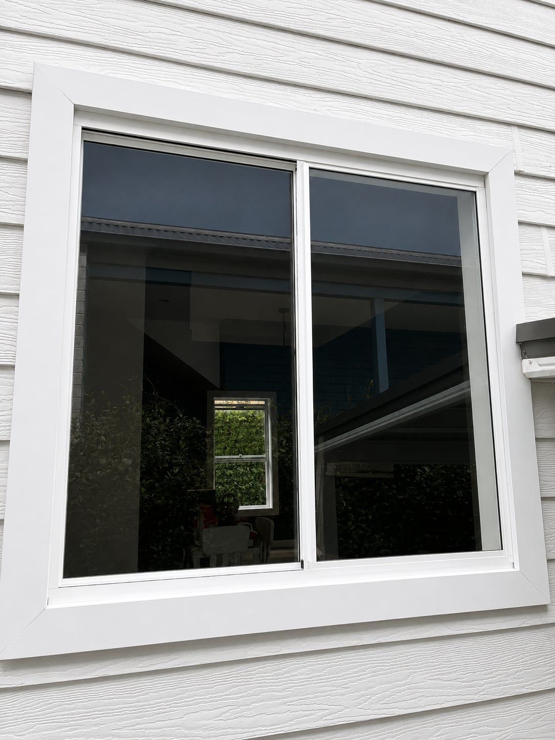 White-framed sliding window in a white exterior wall, reflecting a dark interior and greenery outside — Kracken Windows and Doors In Terrey Hills, NSW