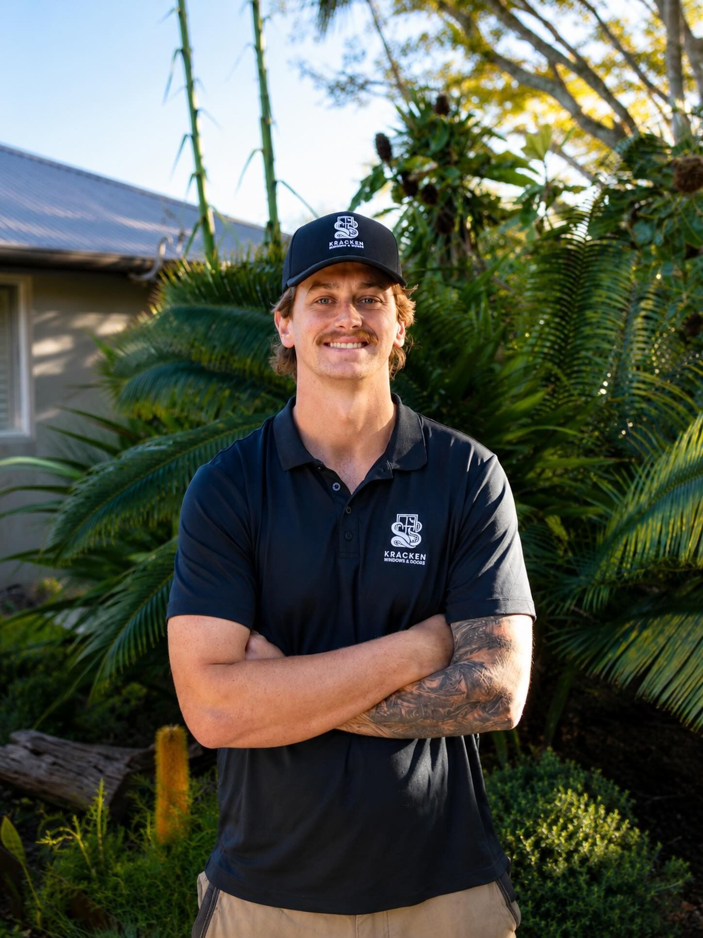 Smiling man in black shirt and cap standing outdoors with tropical plants behind him — Kracken Windows and Doors In Terrey Hills, NSW