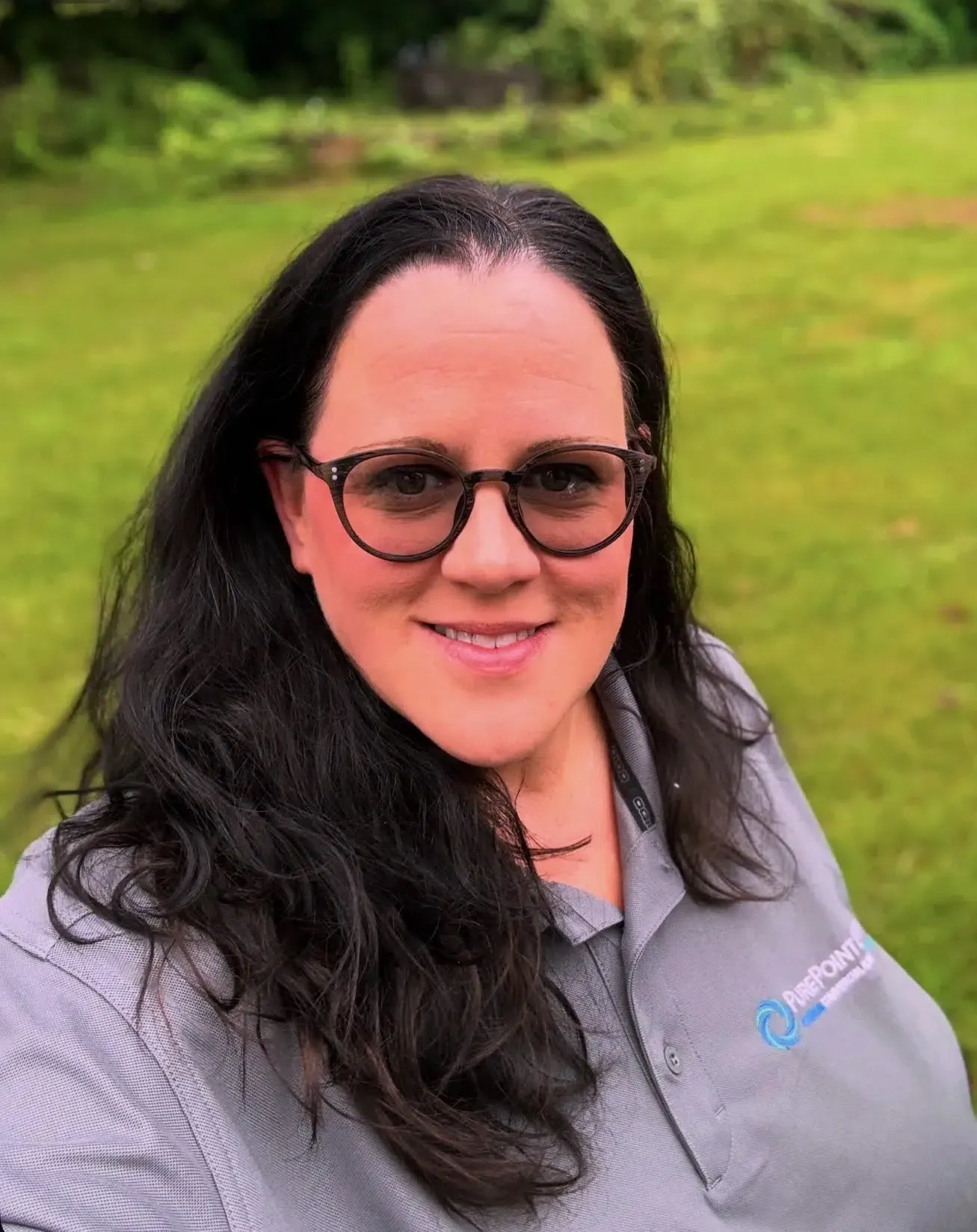 Smiling woman with glasses and dark hair in a gray PurePoint Energy shirt, standing outdoors on a green lawn.