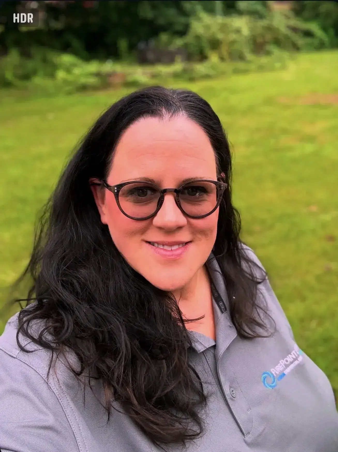Person wearing glasses and a gray shirt, smiling outdoors against a green background.