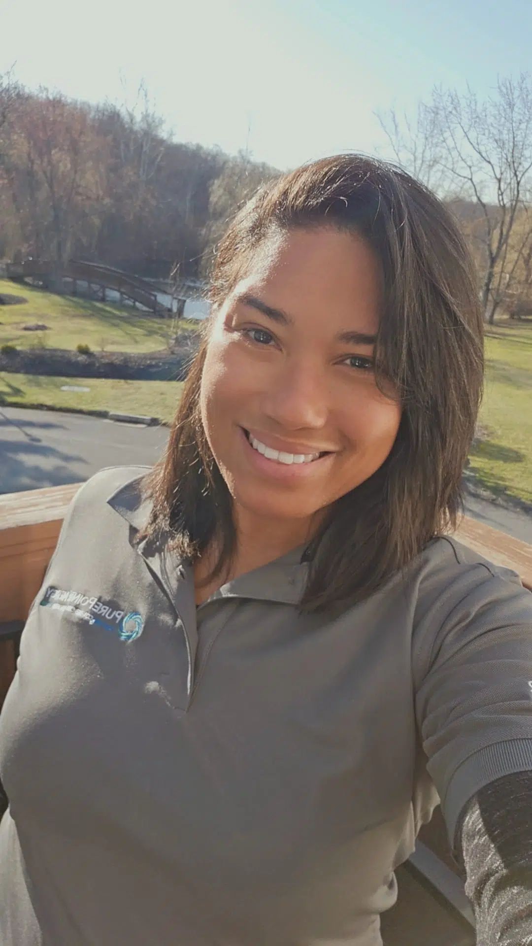 Smiling person taking a selfie outdoors in a sunny park with trees and a lake in the background