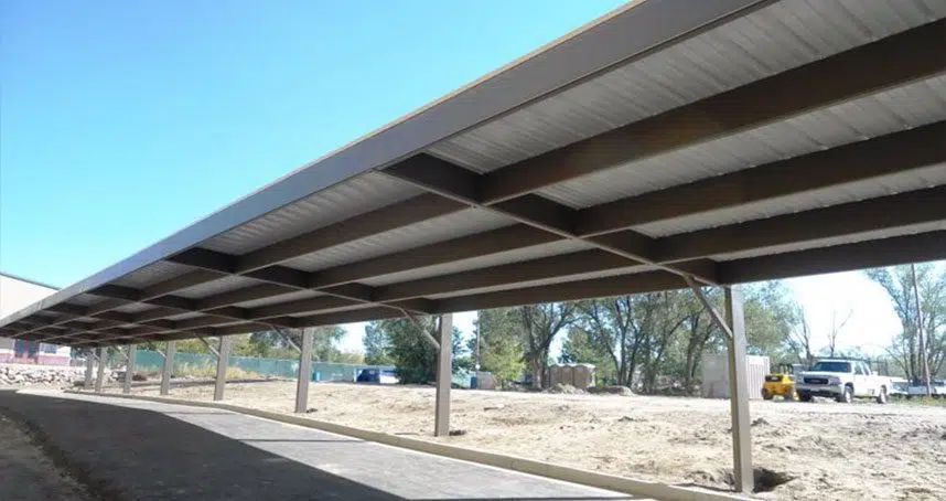 Long metal carport canopy with rows of support posts over a sandy lot under a clear blue sky