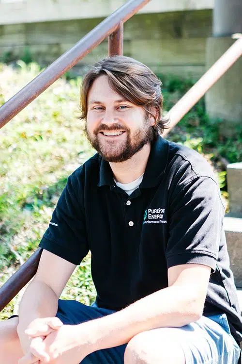 Smiling man in a black PurePoint Energy polo shirt sitting outdoors in sunlight near a railing and greenery