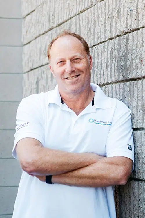 Smiling man in a white PurePoint Energy polo shirt stands with arms crossed beside a stone wall.