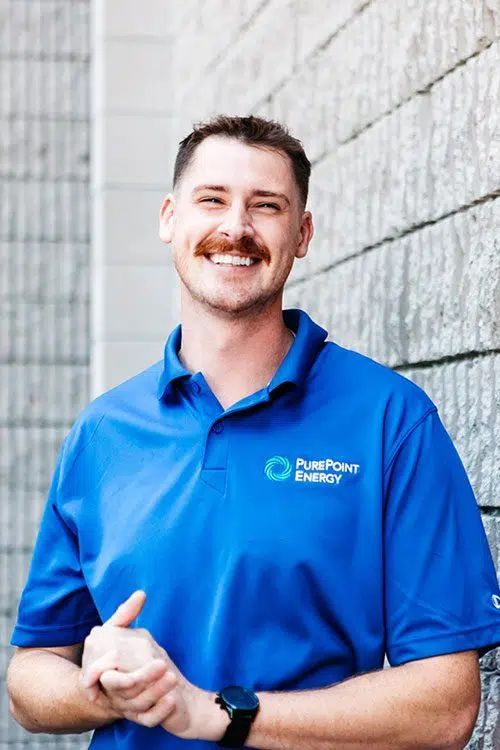 Smiling man in blue PurePoint Energy polo shirt standing against a light gray stone wall.