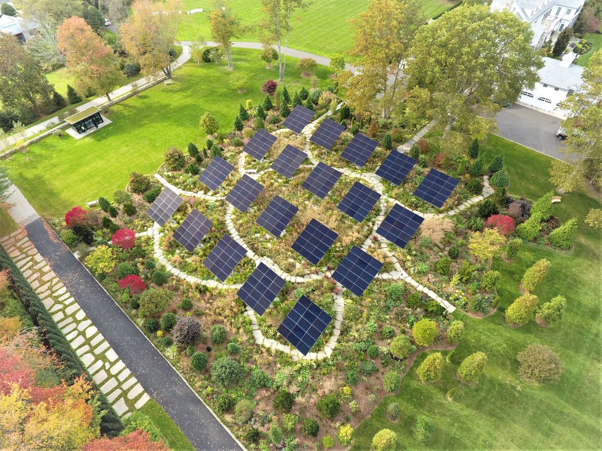 Aerial view of a garden-like solar farm with rows of blue solar panels surrounded by trees and flowers
