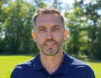 Man standing outdoors in a navy PurePoint Energy polo shirt with trees and a sunny green background.