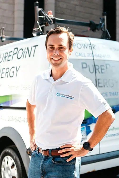 Man in white polo shirt standing in front of a PurePoint Energy service van, smiling with hands on hips