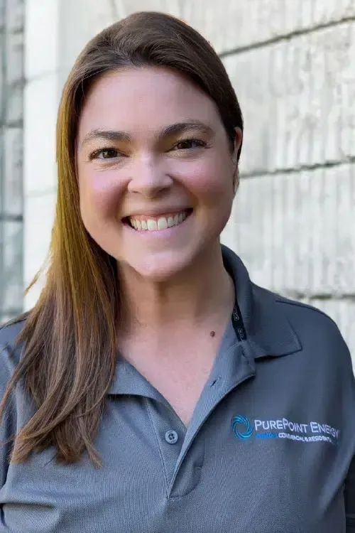 Smiling person in a gray PurePoint Energy polo shirt with a logo, standing outdoors by a light brick wall