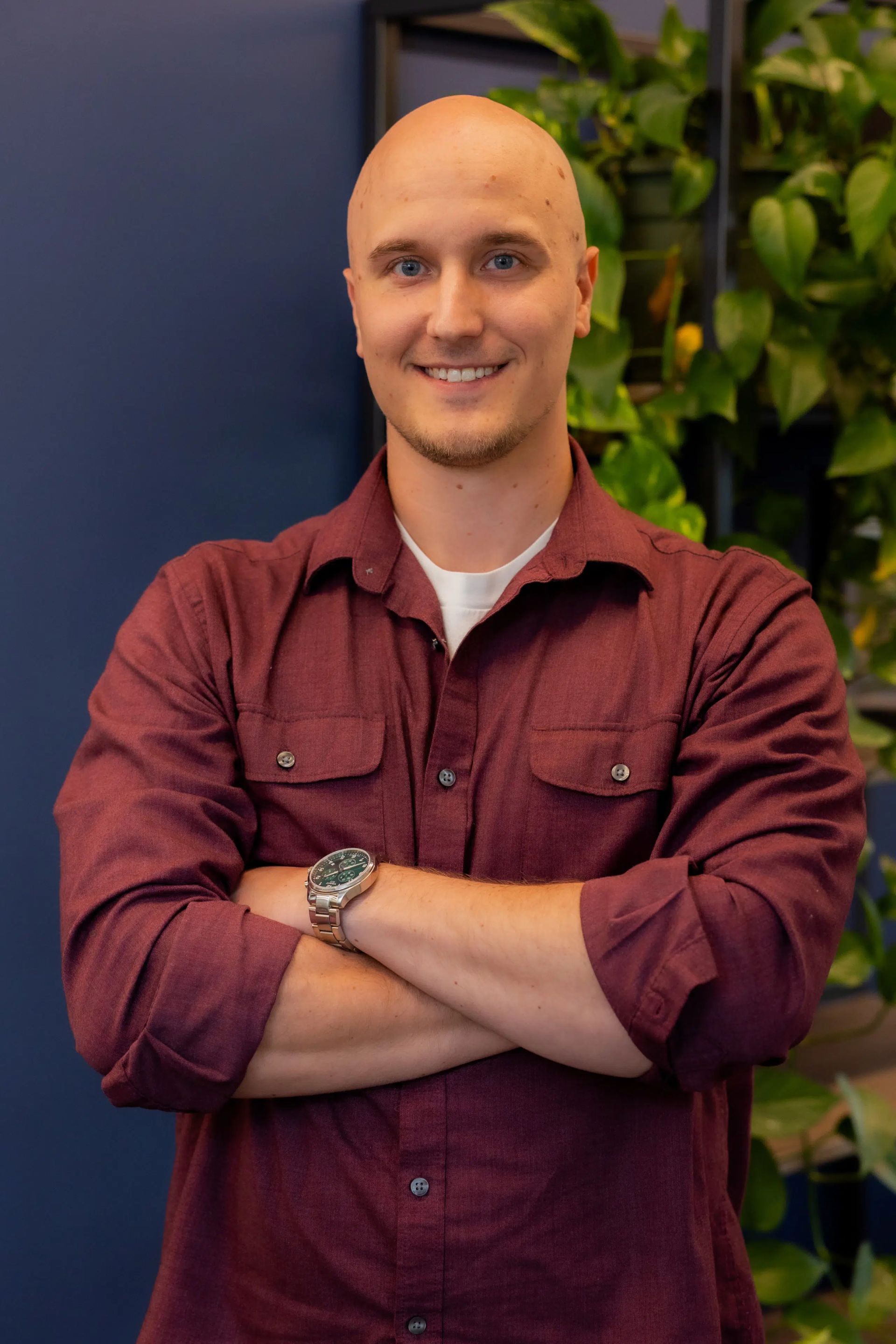 Smiling bald man in a maroon PurePoint Energy shirt with folded arms, standing beside a leafy green wall.