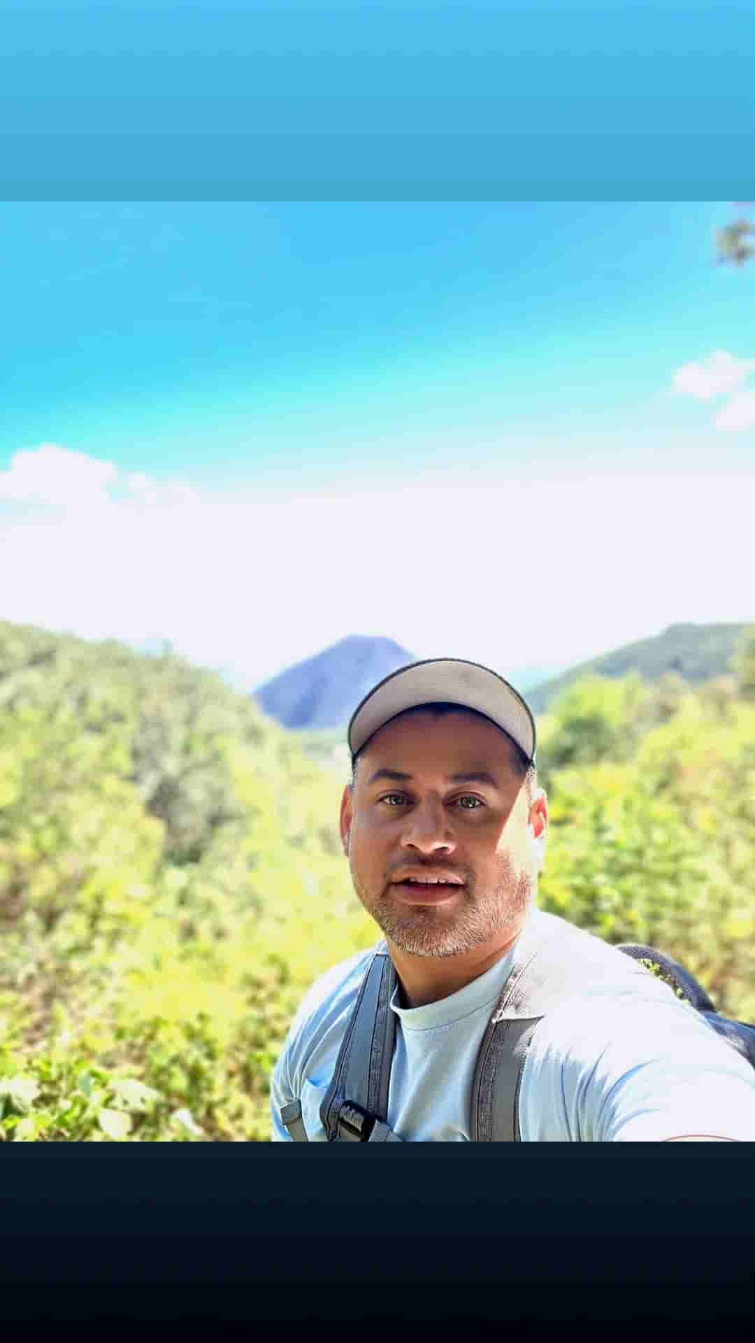 Man taking a selfie outdoors with bright blue sky, green hills, and sunlight behind him