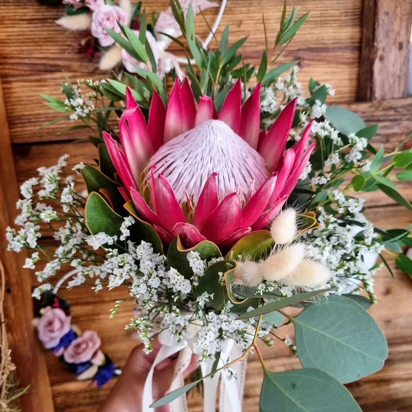 Bouquet di protee rosa con fiori di riempimento bianchi e verde su una superficie di legno.