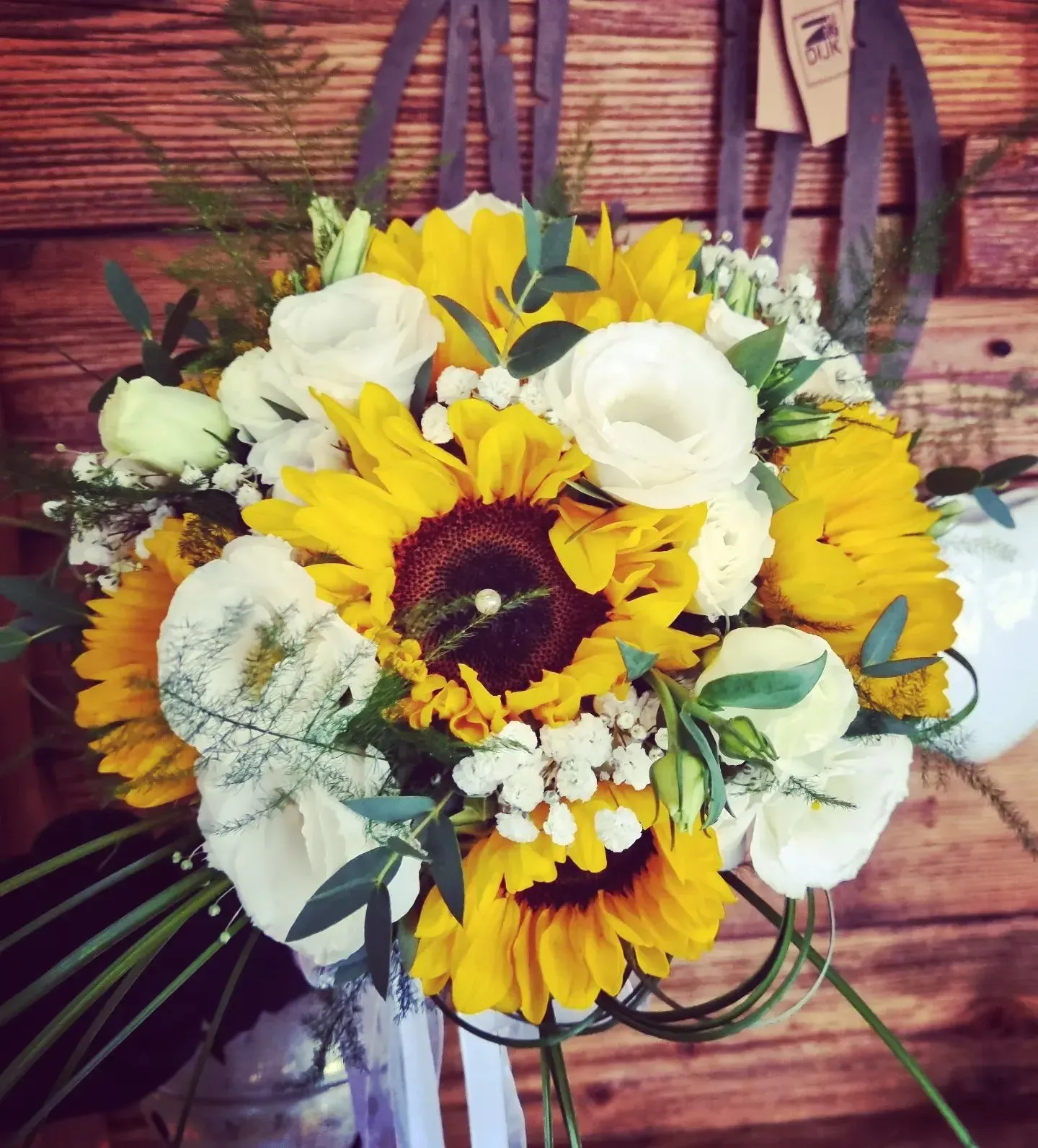 Bouquet di girasoli e rose bianche con verde in un cestino marrone su uno sfondo di legno rustico.
