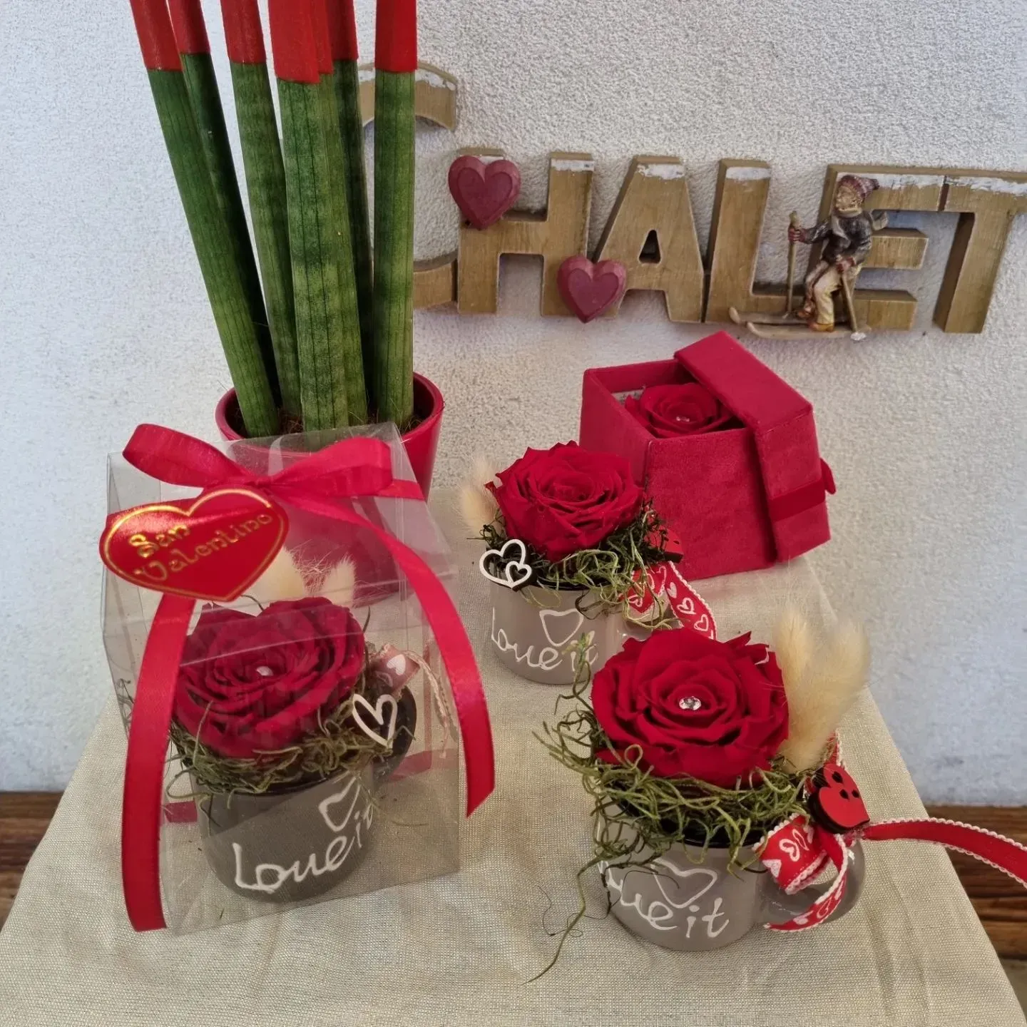 Due composizioni di rose rosse con nastri e una pianta di rose in un vaso di vetro accanto alle lettere decorative 