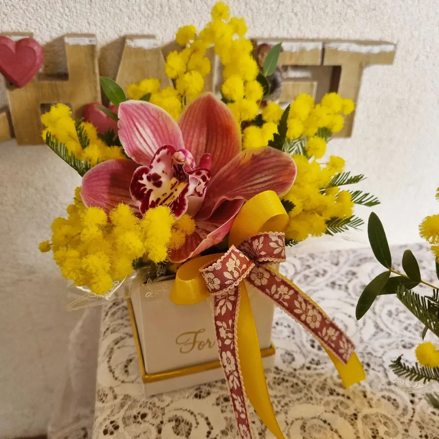 Orchidea rosa e fiori gialli in una confezione regalo decorata con un nastro, su una tovaglia di pizzo.