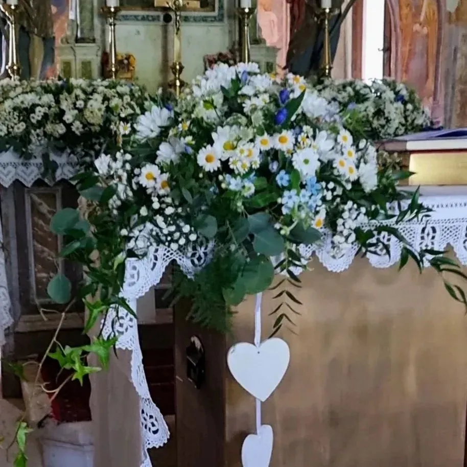 Composizione floreale bianca per l'altare, con drappeggi di pizzo e decorazioni a forma di cuore pendenti, in un ambiente ecclesiastico.