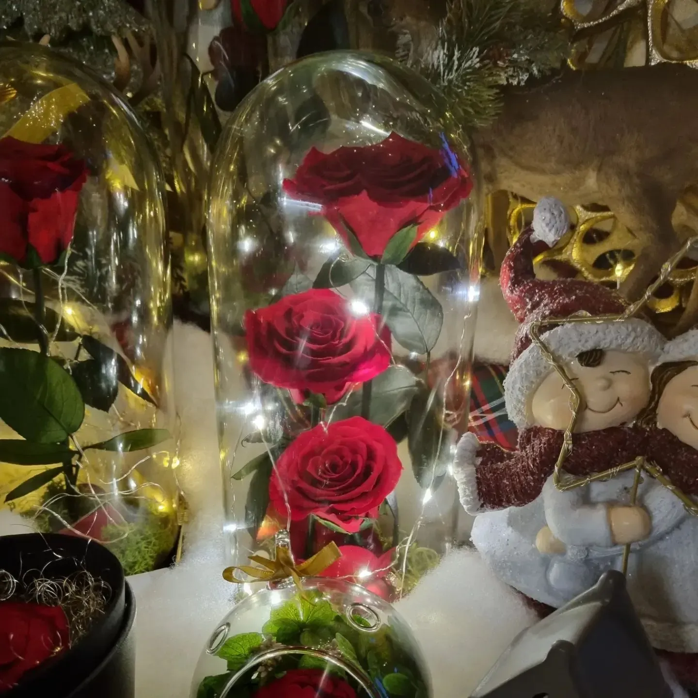 Cupole di vetro con rose rosse e un orsacchiotto in una scenografia natalizia innevata, illuminate da calde lucine.