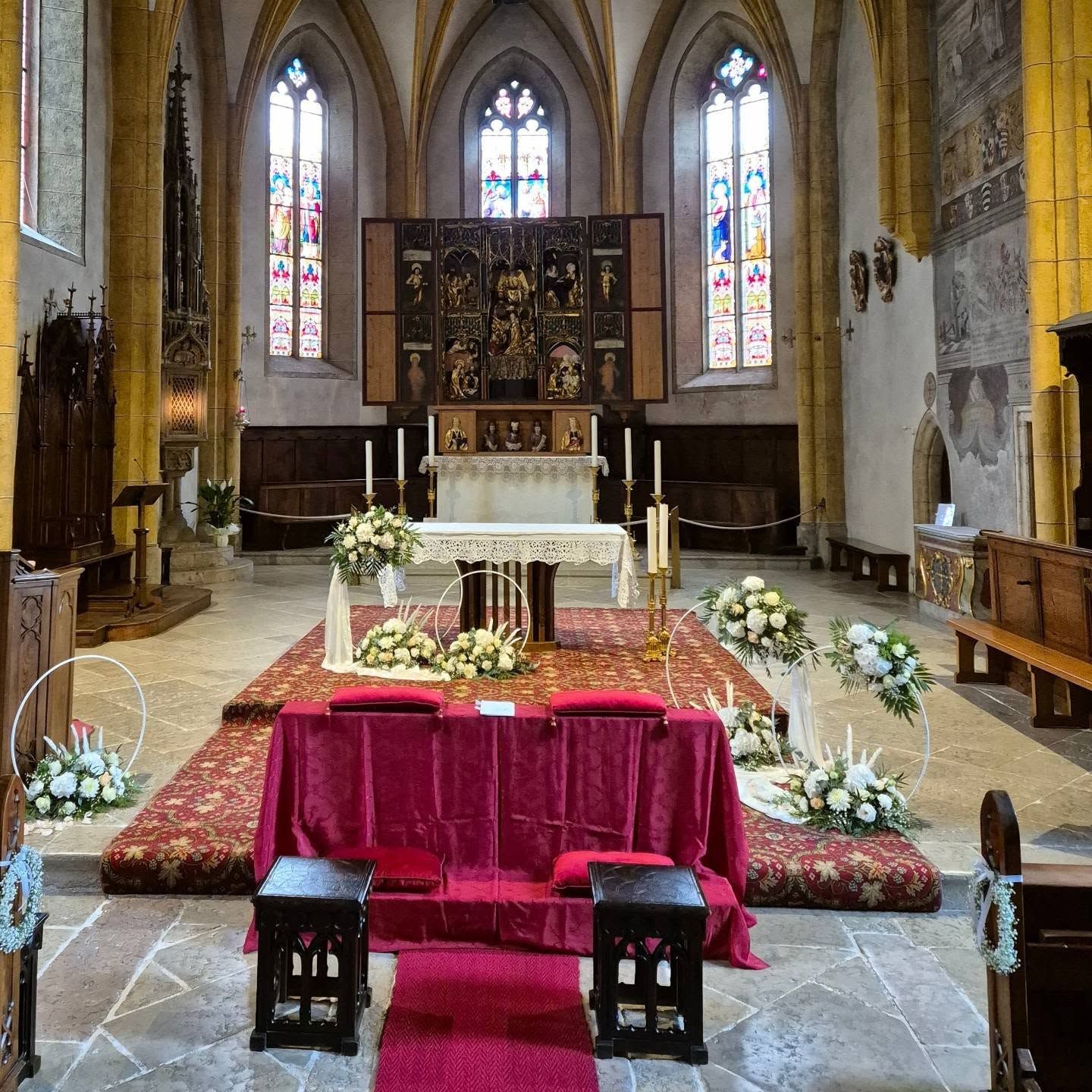 Altare della chiesa con tovaglia rossa, fiori, candele e vetrate colorate sullo sfondo.