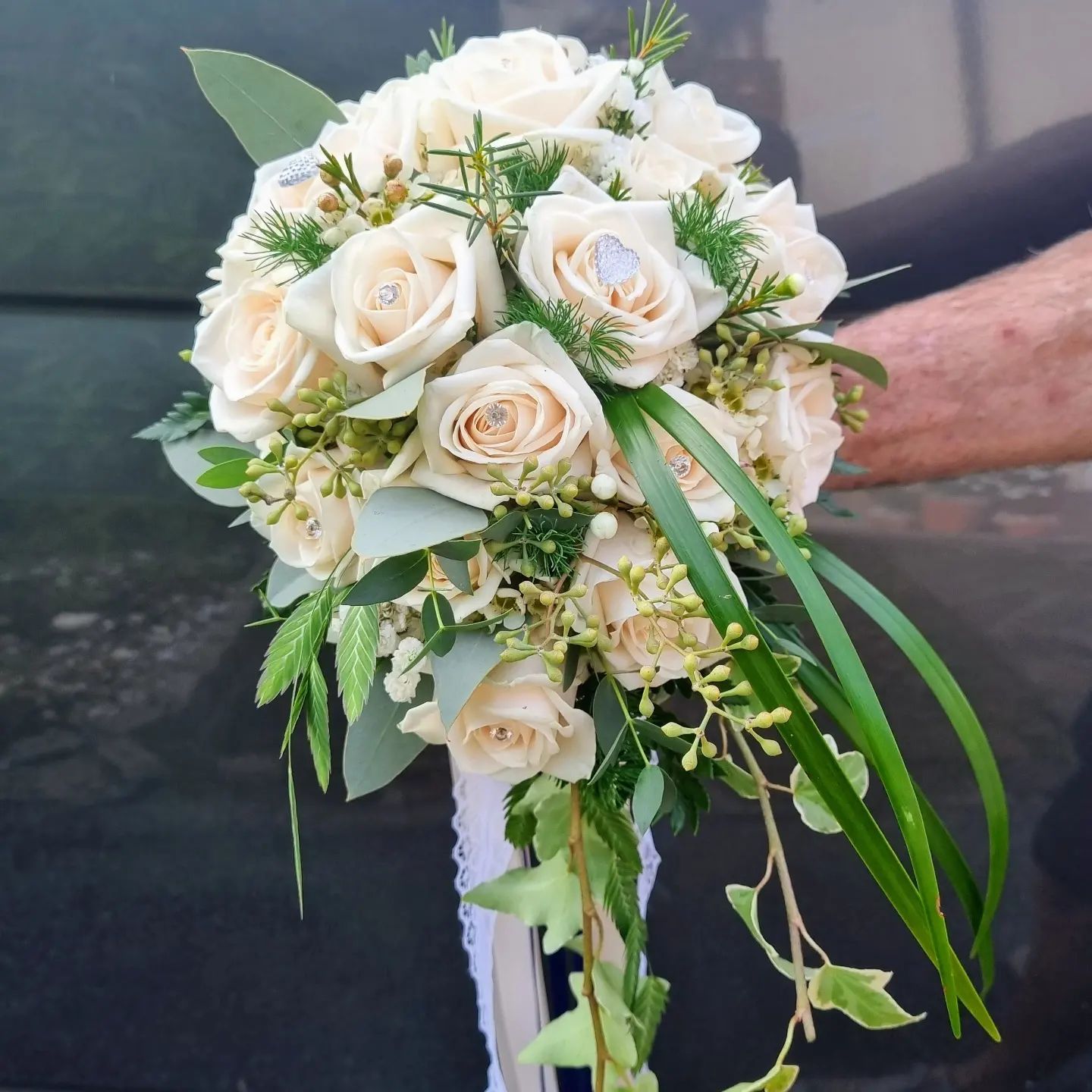 Bouquet di rose color crema con fiori bianchi, fogliame ricadente e lunghi nastri decorativi, tenuto in mano.