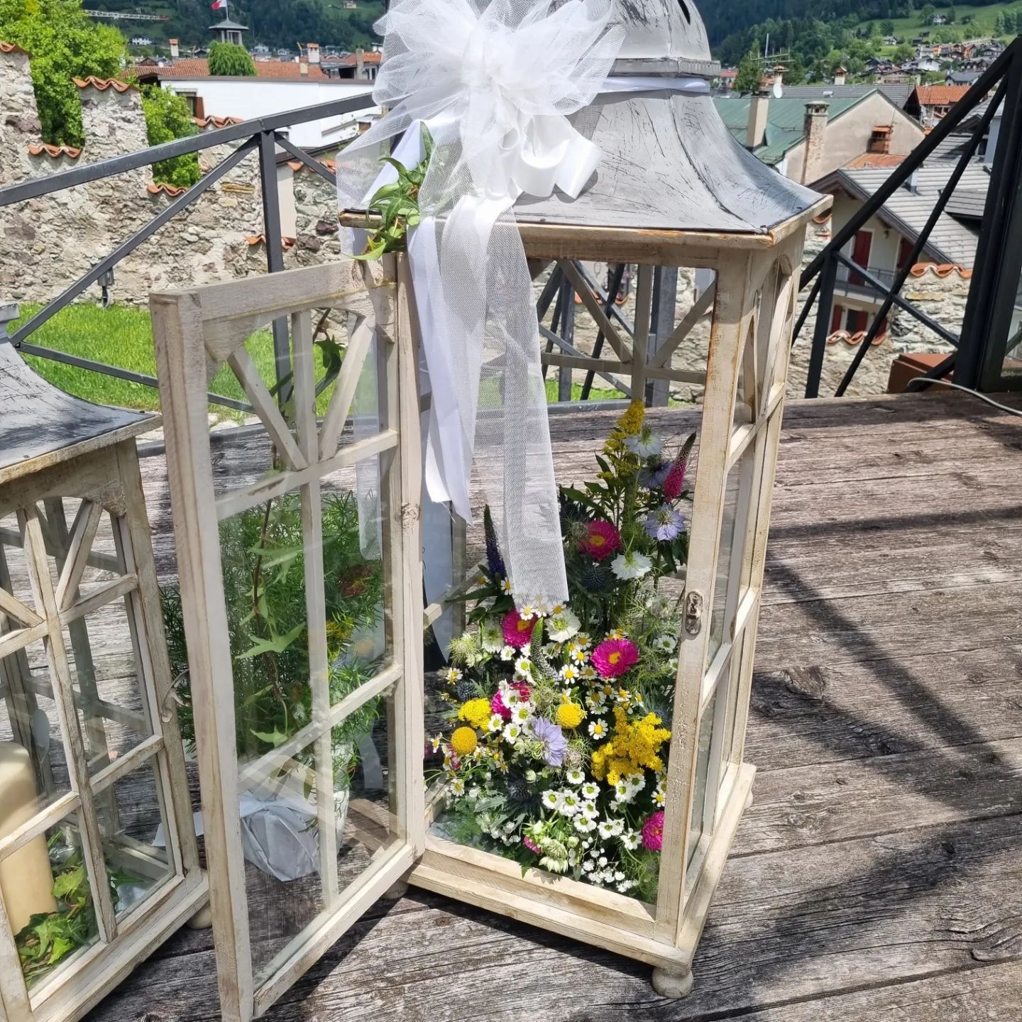 Lanterna bianca con nastro e fiori su una terrazza di legno, affacciata su un cimitero.