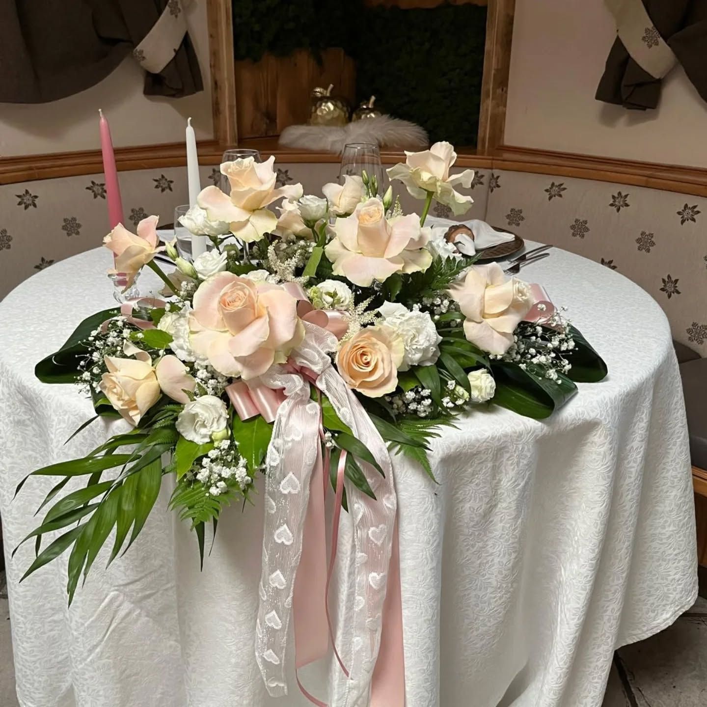 Elegante centrotavola floreale con rose bianche e rosa cipria e nastri rosa su un tavolo rotondo in sala da pranzo.