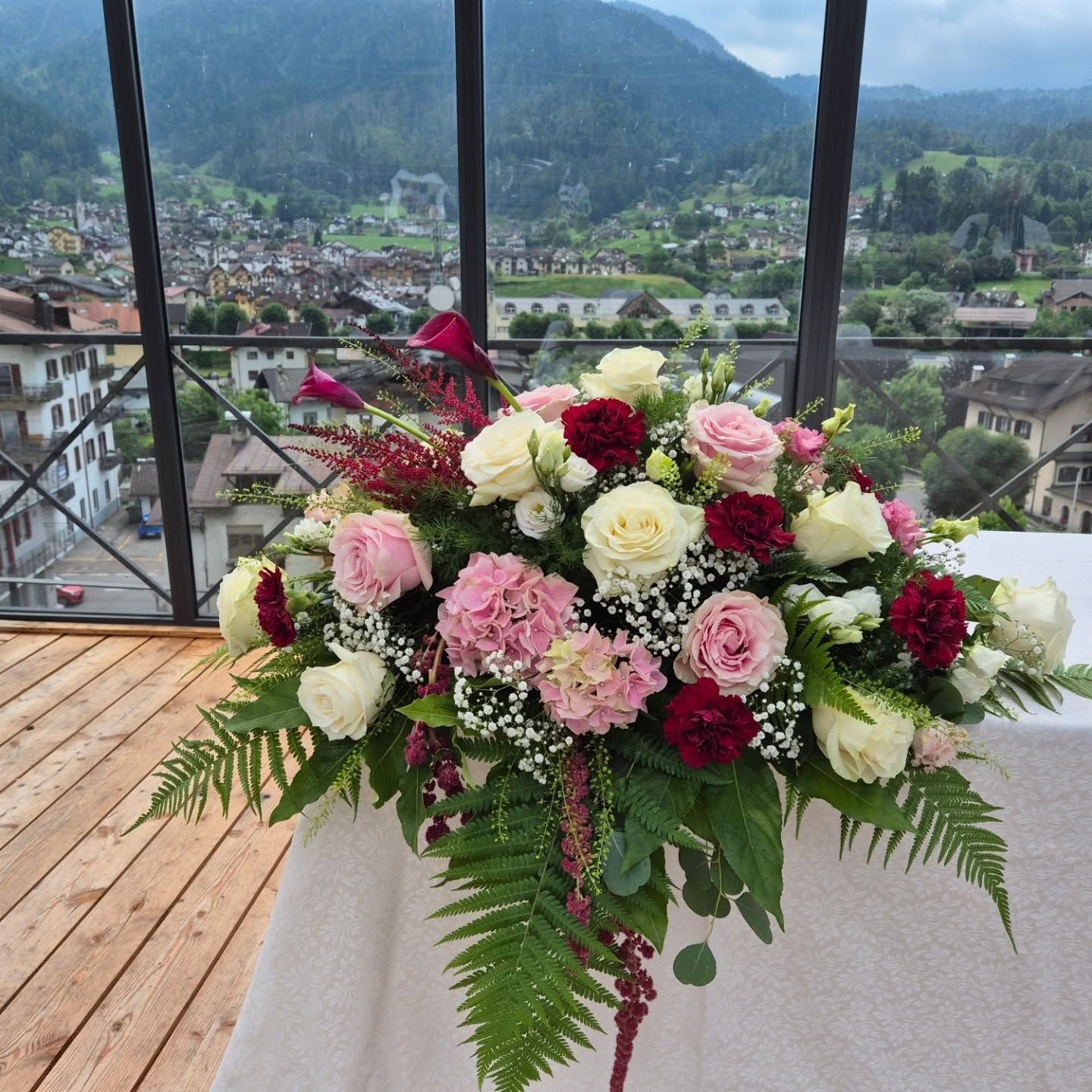 Composizione floreale rosa, bianca e bordeaux su una terrazza con vista sulle montagne e sul villaggio sullo sfondo.