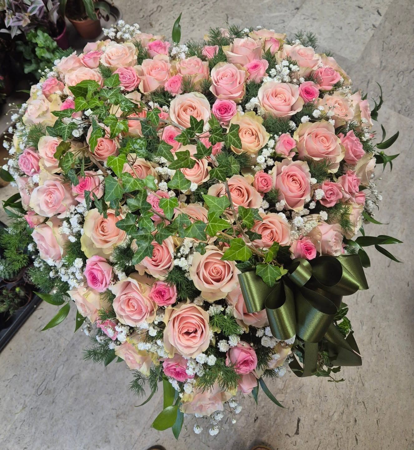 Grande bouquet a forma di cuore composto da rose rosa e fiori bianchi con verde, impreziosito da un nastro scuro, su una superficie di pietra.