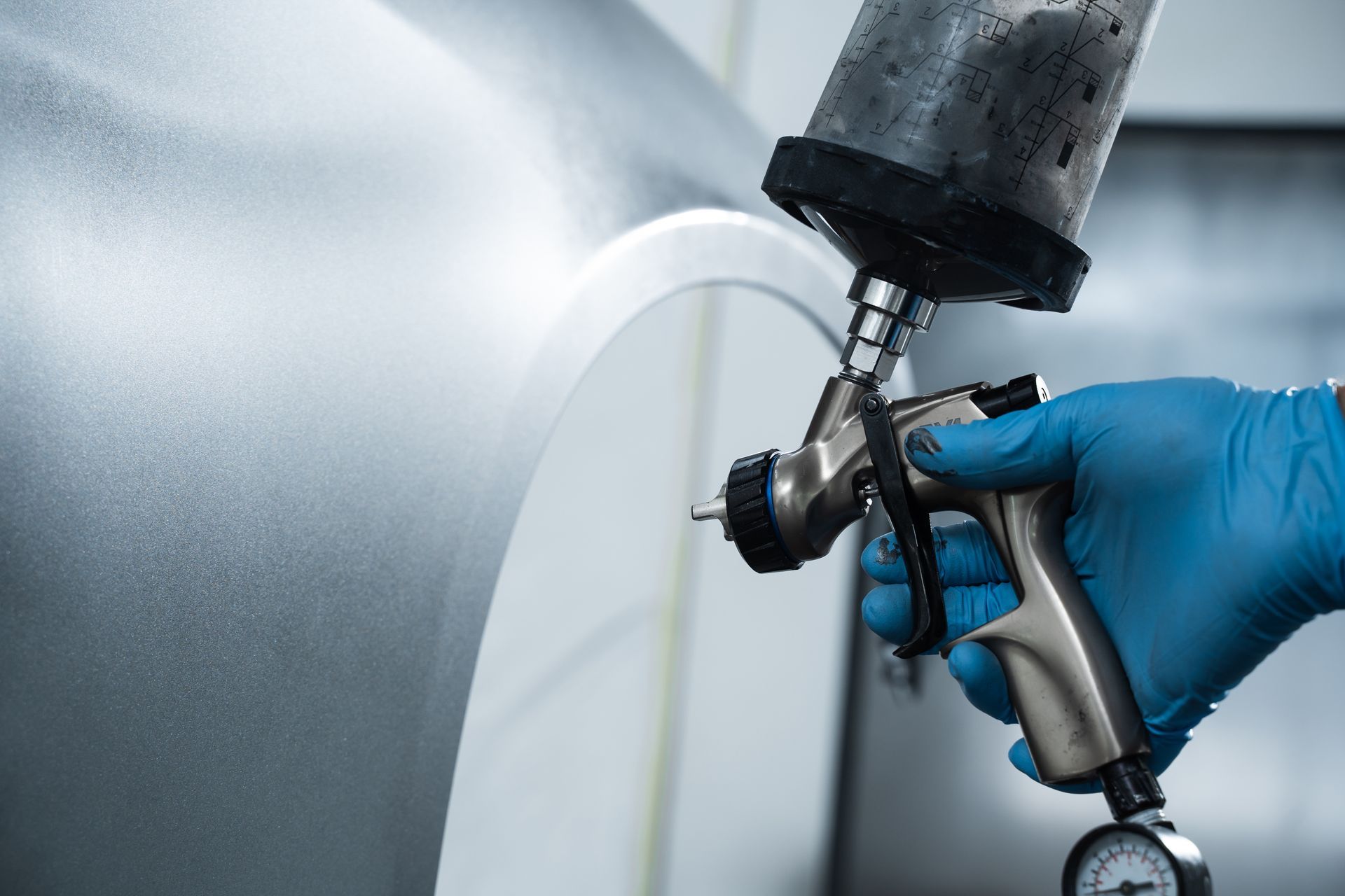 Person in blue gloves using a spray gun to paint a car panel silver in a workshop. Person in blue gloves using a spray gun to paint a car panel silver in a workshop.